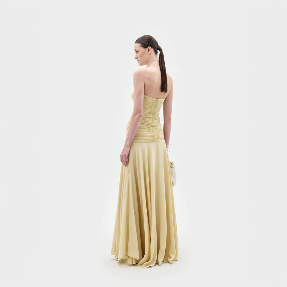 Una mujer de espaldas luce el Norma Dress, un maxi vestido strapless de silueta columna en amarillo claro. Lleva el cabello castaño y liso en una coleta baja y sostiene un bolso blanco. Fondo blanco liso.