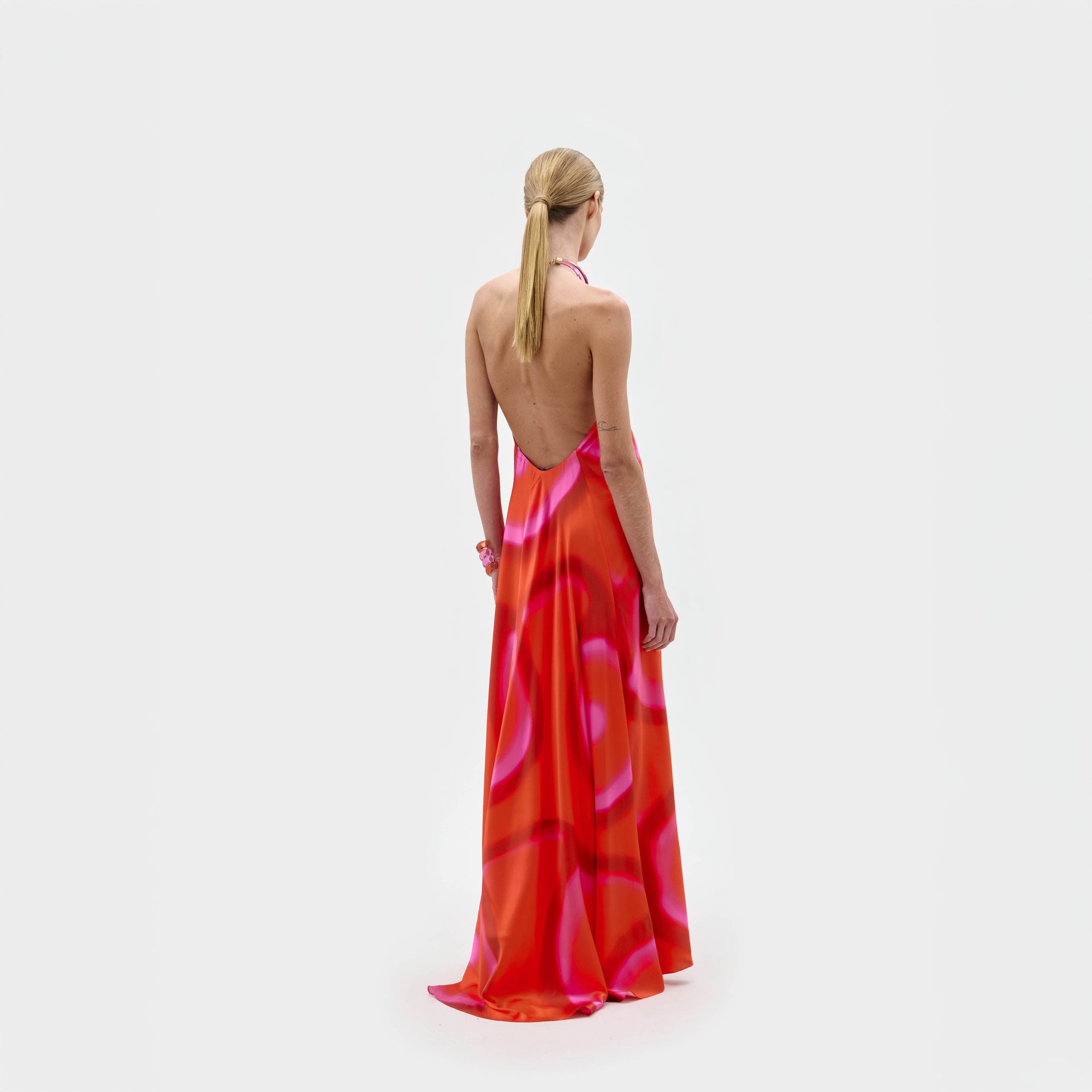 Una persona con el pelo rubio recogido en una coleta baja, de espaldas a la cámara, luce el Jesse Dress, un vestido maxi con un estampado abstracto rojo y rosa, sobre un fondo blanco liso. Pieza exclusiva fabricada en Colombia.