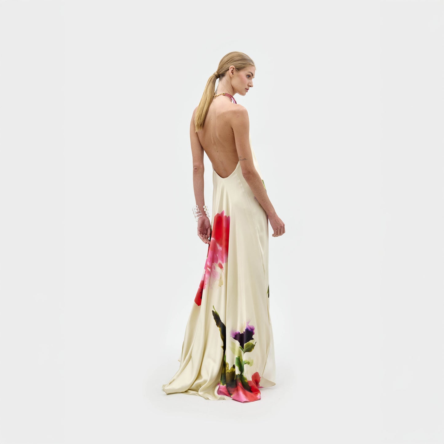 Una mujer de pelo largo y rubio lleva el vestido Jesse, un maxi vestido de seda color crema con escote halter, espalda abierta y llamativos estampados florales en el bajo. Está de espaldas, mirando por encima del hombro sobre un fondo blanco liso. Hecho en Colombia.