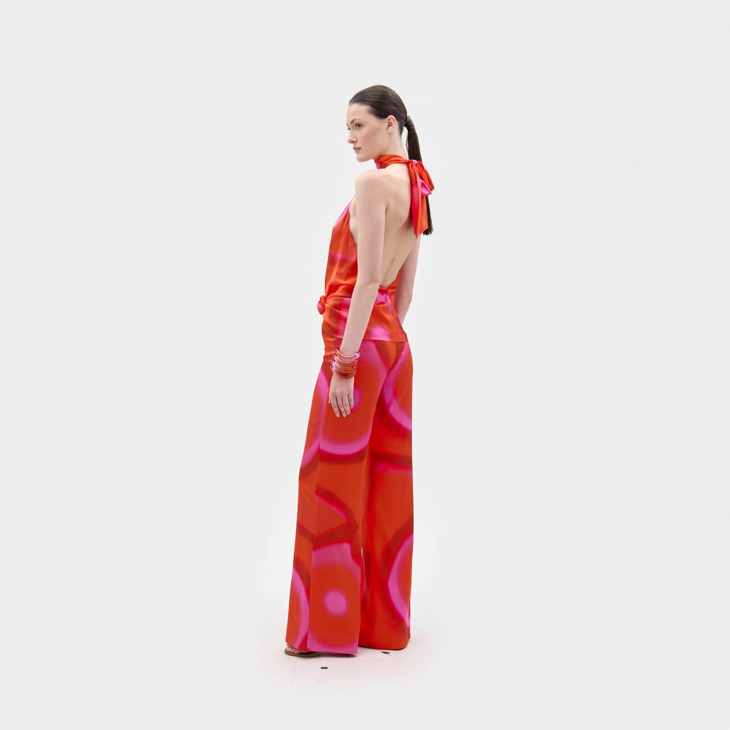 Una persona, de pie sobre un fondo blanco liso, lleva puesto el Andie Pant, un mono rojo sin espalda, con cuello halter, piernas anchas y un estampado abstracto rosa. La persona mira de reojo hacia la cámara.