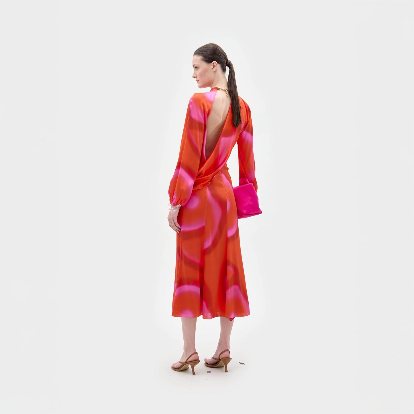 Una mujer mira hacia otro lado con el vestido Muna, una cintura entallada midi con brillantes estampados naranjas y rosas en lustrosa seda italiana. Lo combina con un bolso de mano rosa a juego, sandalias de tacón color canela y el pelo recogido en una coleta baja.