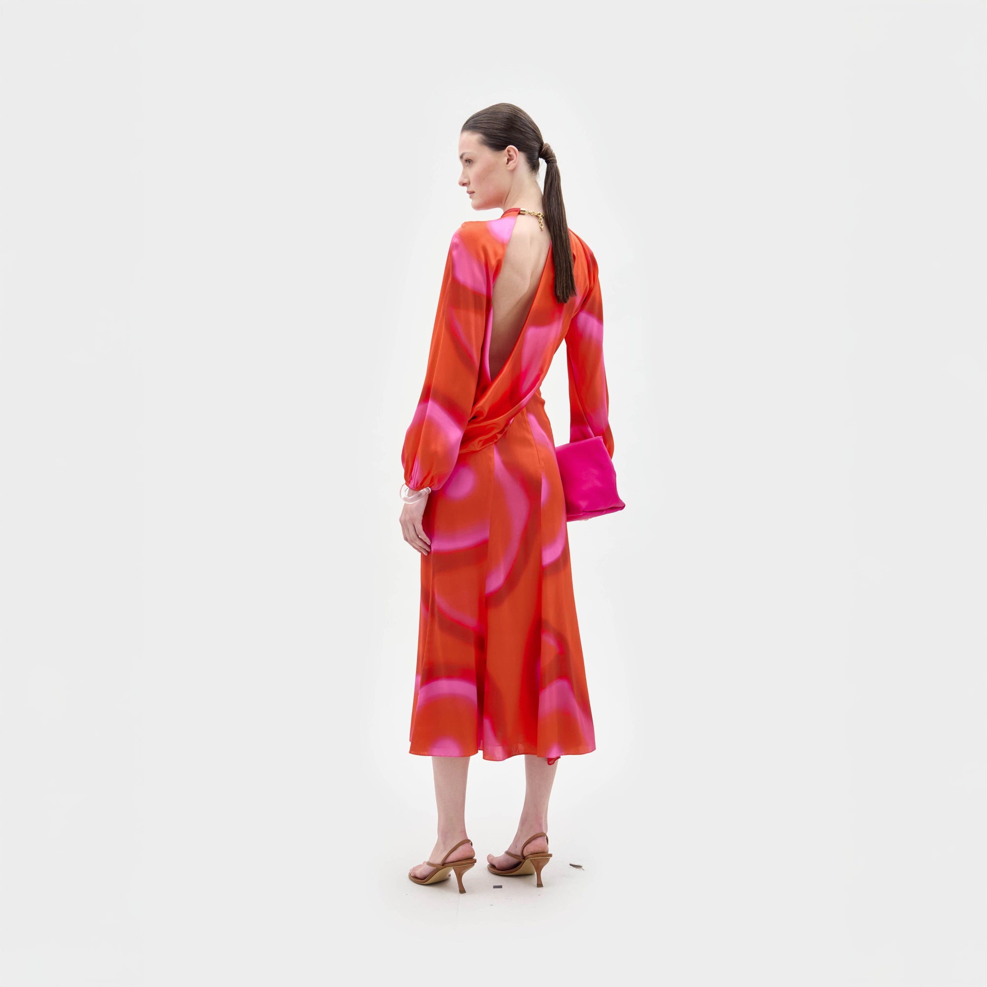 Una mujer mira hacia otro lado con el vestido Muna, una cintura entallada midi con brillantes estampados naranjas y rosas en lustrosa seda italiana. Lo combina con un bolso de mano rosa a juego, sandalias de tacón color canela y el pelo recogido en una coleta baja.