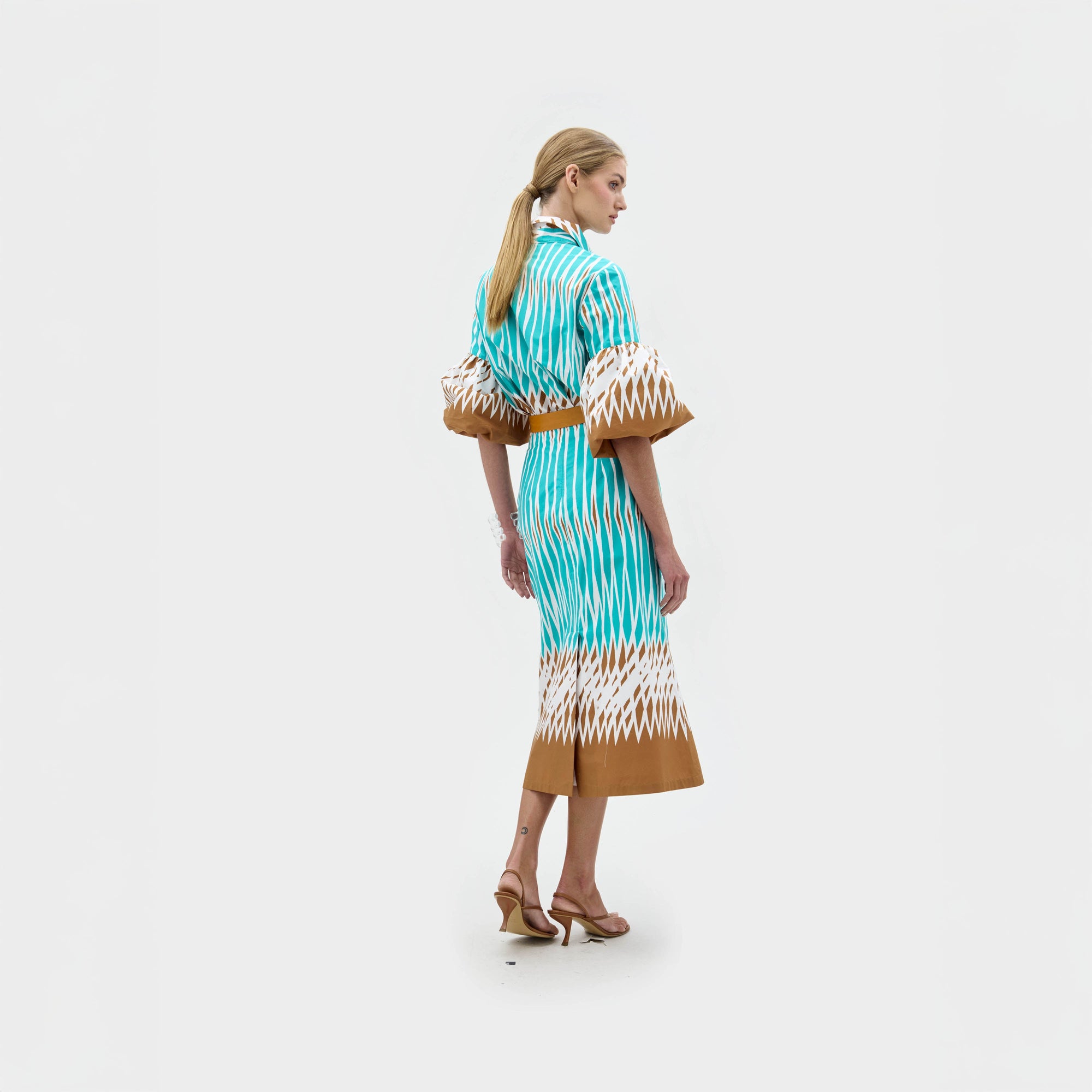Sobre un fondo blanco, una mujer con el pelo claro recogido en una coleta baja lleva la falda Atira, una falda midi de talle alto con estampado turquesa y marrón, combinada con mangas acampanadas y sandalias de tacón beige.