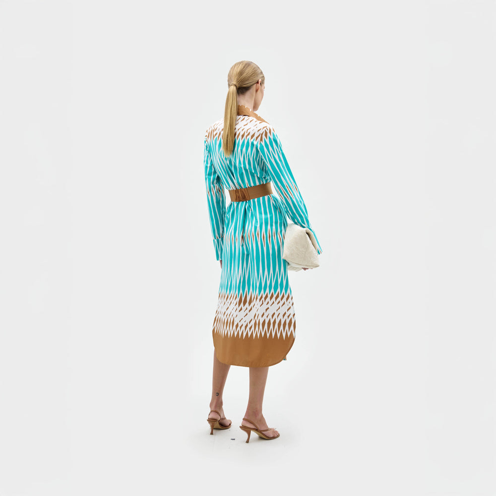 Una mujer mira hacia otro lado y lleva el vestido Ronzona con motivos azules, blancos y marrones y un cinturón marrón. Lleva un bolso de mano blanco, sandalias de tacón marrones y el pelo rubio recogido en una coleta baja. Hecho en Colombia.