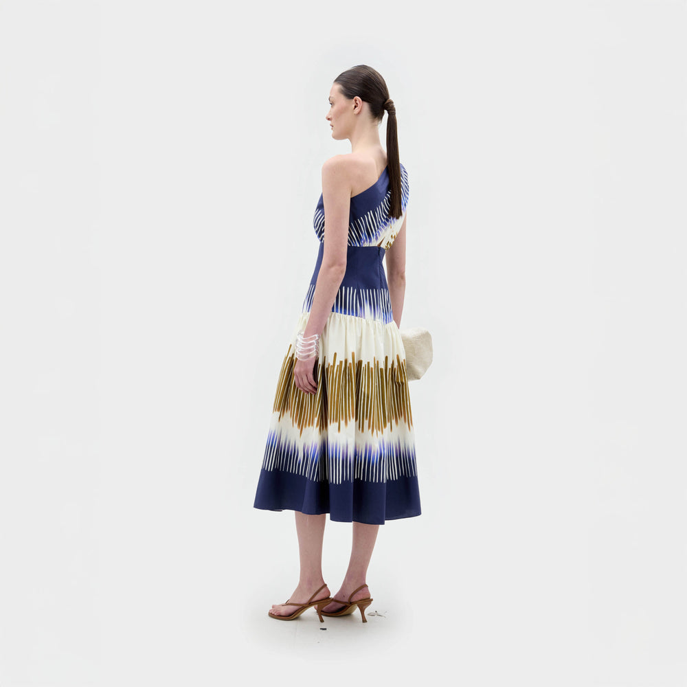 Una mujer sobre un fondo blanco liso lleva el vestido Catalina, con estampado azul, blanco y dorado y falda midi acampanada. Lleva el pelo castaño recogido en una coleta baja y sandalias de tacón.