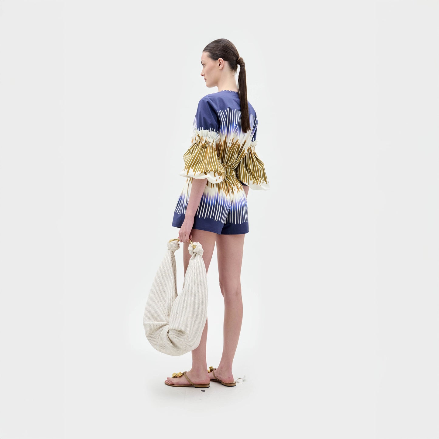 Una mujer está de pie sobre un fondo blanco, mirando ligeramente hacia otro lado. Lleva el Giorgio Short -un conjunto corto de talle alto con estampado azul y blanco y mangas con flecos dorados-, sandalias planas y un bolso grande de color hueso en la mano izquierda.