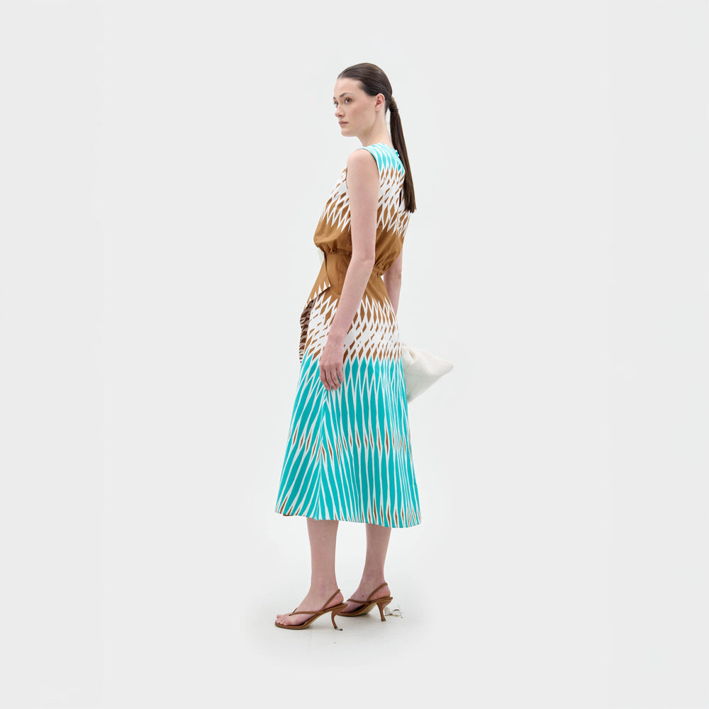 Una mujer, de perfil sobre un fondo blanco liso, lleva el vestido Kendall Dress -un vestido midi ajustado con estampado geométrico en marrón y azul- combinado con sandalias de tacón marrones, un bolso de mano blanco y el pelo oscuro recogido en una coleta baja.