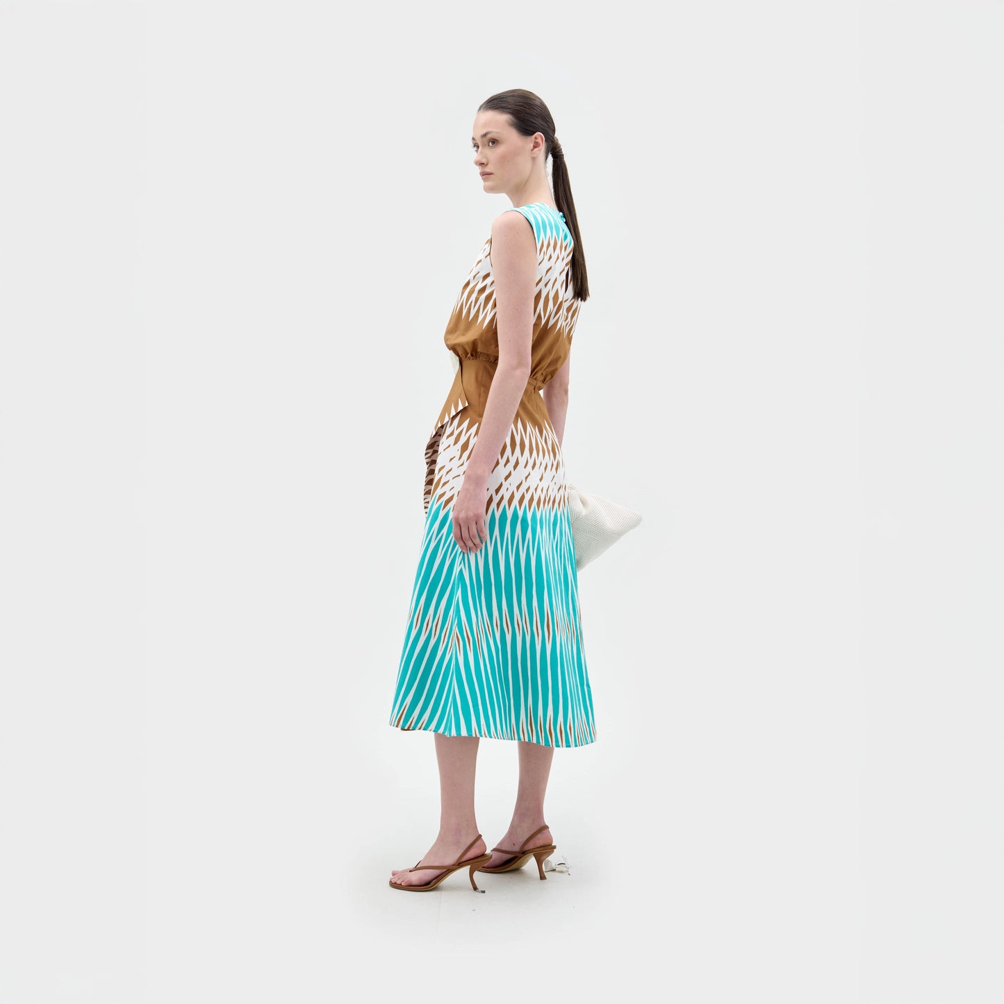Una mujer, de perfil sobre un fondo blanco liso, lleva el vestido Kendall Dress -un vestido midi ajustado con estampado geométrico en marrón y azul- combinado con sandalias de tacón marrones, un bolso de mano blanco y el pelo oscuro recogido en una coleta baja.
