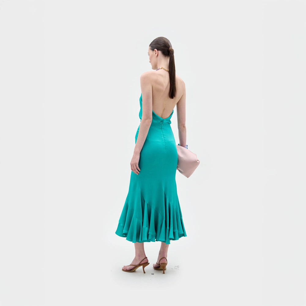 Sobre un fondo blanco liso, una mujer lleva la falda Marlene -un vestido verde azulado con escote halter y espalda descubierta, fabricado en Colombia- y sandalias de tacón marrones. Lleva un bolso de mano rosa pálido y el pelo liso y castaño recogido en una coleta.