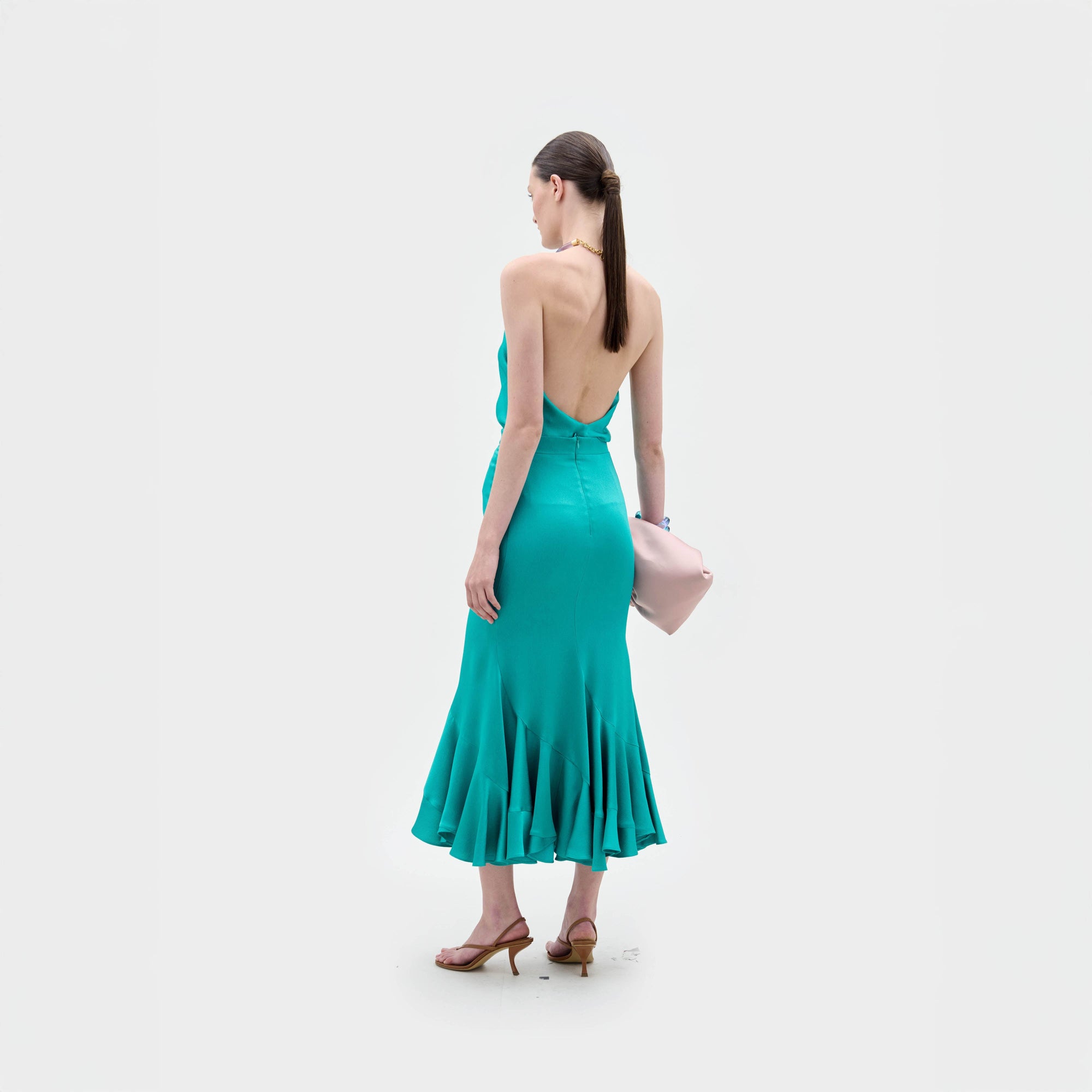 Sobre un fondo blanco liso, una mujer lleva la falda Marlene -un vestido verde azulado con escote halter y espalda descubierta, fabricado en Colombia- y sandalias de tacón marrones. Lleva un bolso de mano rosa pálido y el pelo liso y castaño recogido en una coleta.