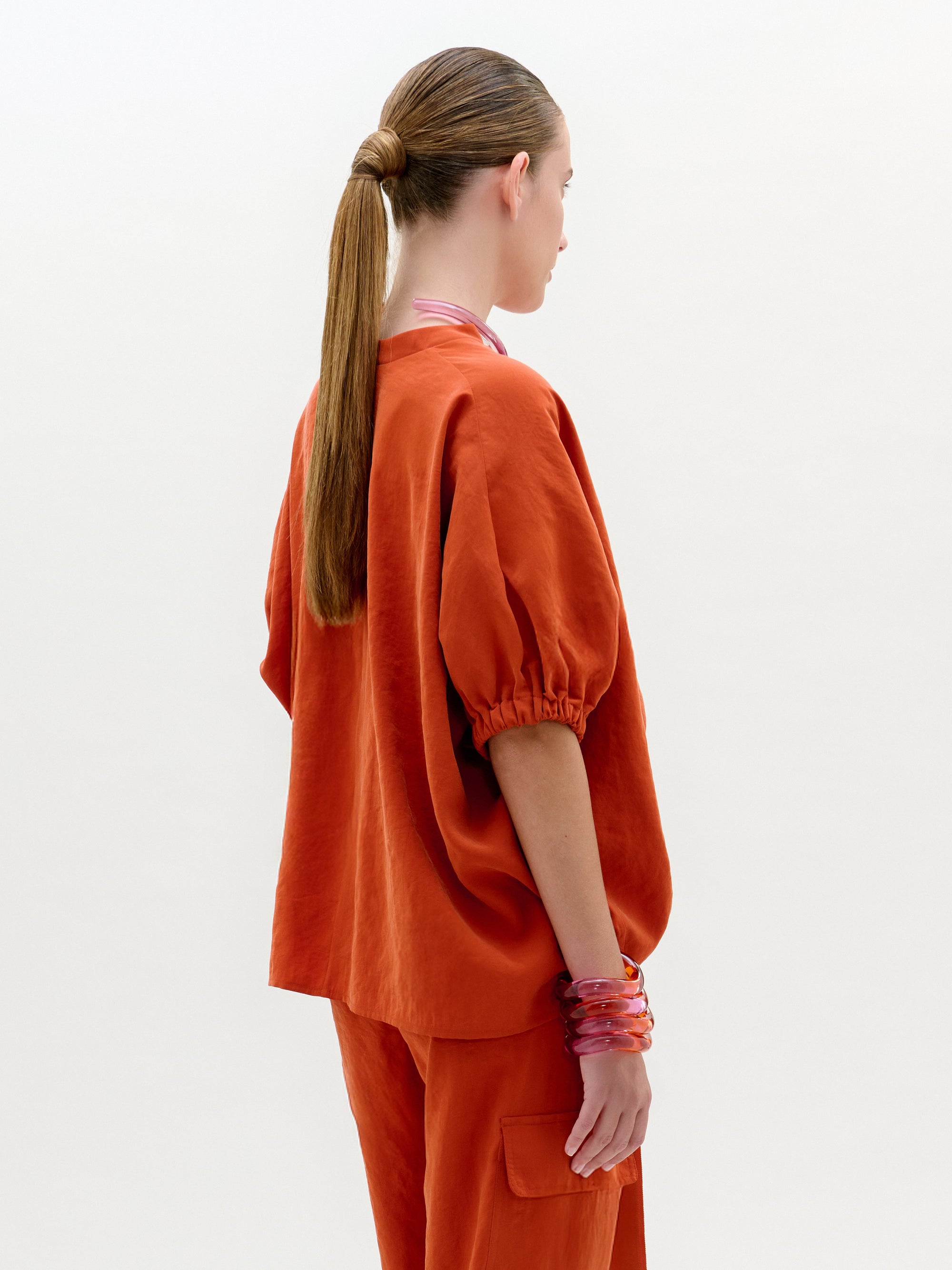 Una persona con el pelo largo y liso recogido en una coleta baja se ve de perfil y de espaldas, llevando la Blusa Susane -un top naranja de manga corta y pantalones a juego- con varias pulseras rosas en la muñeca izquierda. Fabricada en Colombia.