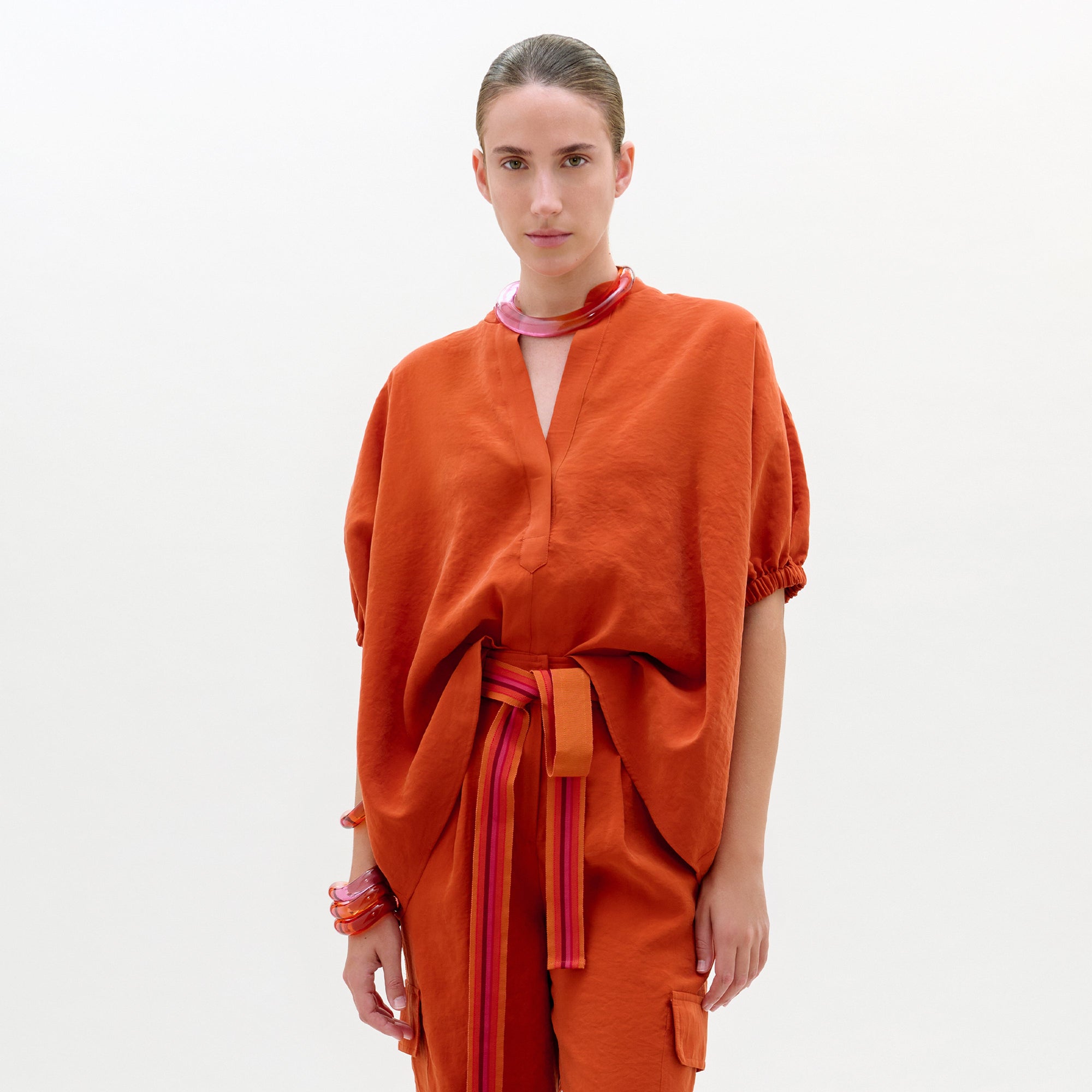 Una persona, sobre un fondo claro, lleva la blusa Susane, una blusa naranja quemado de manga corta y cuello en V, con mangas sueltas y un cinturón a rayas rosa-naranja, confeccionada en Colombia con tejido italiano, que se combina con pulseras y un collar grueso.