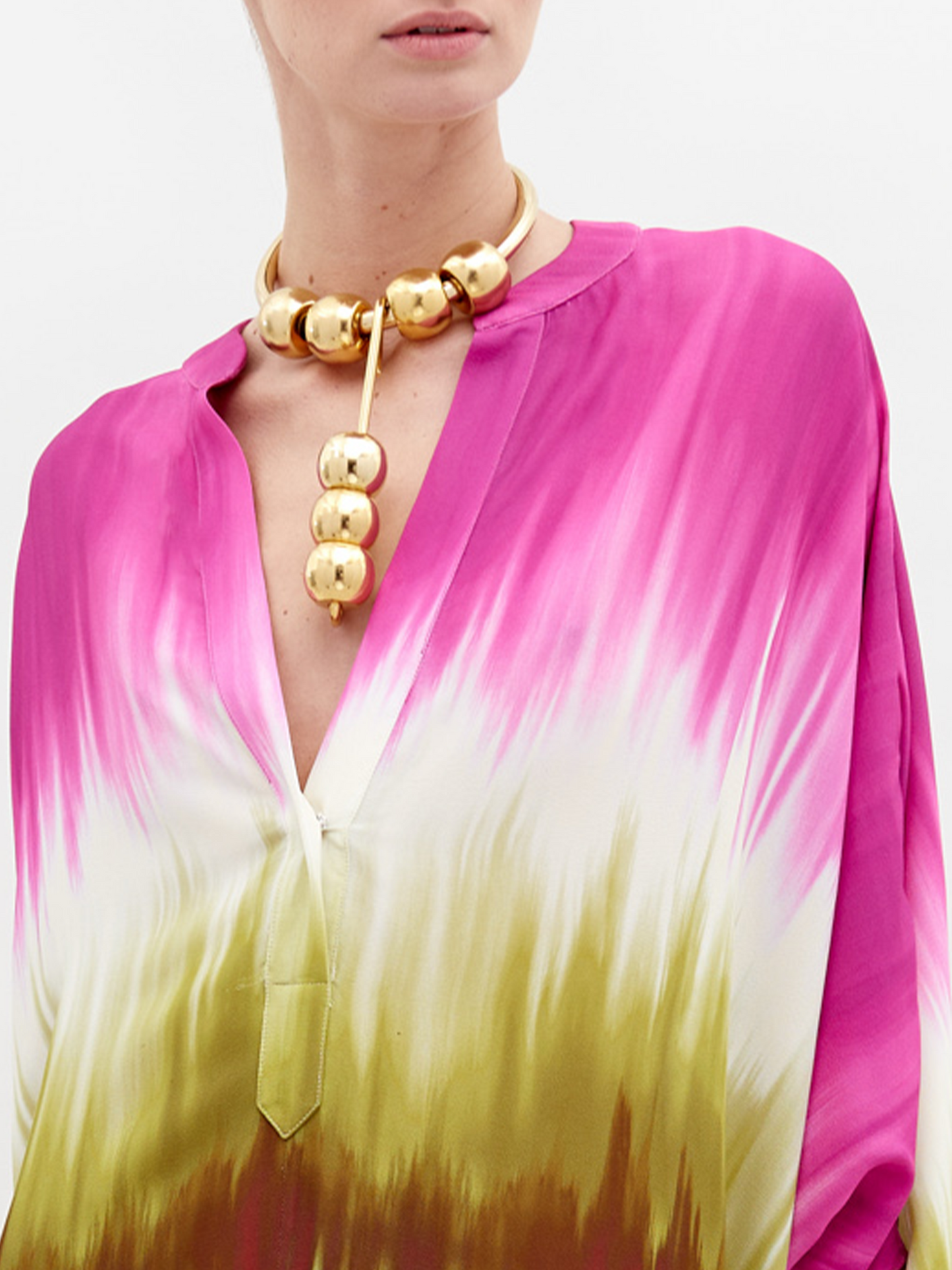 A person is wearing a colorful Serena Tunic Violeta Lime Tie Dye with shades of pink, green, and white. They are accessorized with a chunky gold necklace and sandals. The background is plain white. Get ready to pre-order this look by November 15th, 2024 to ensure you receive it by the ship-by date.