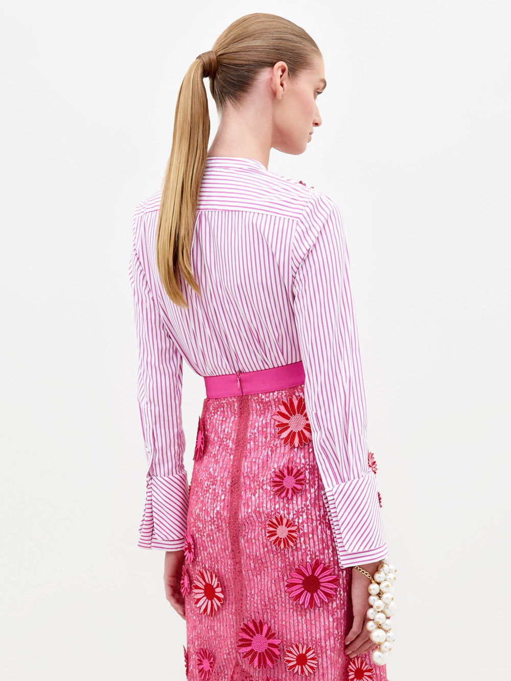 A person in the Silvina Blouse Rose Stripes adorned with floral appliqués, paired with a pink sequined skirt featuring similar floral designs, stands against a plain white background, available for pre-order now with an expected ship date in early 2025.