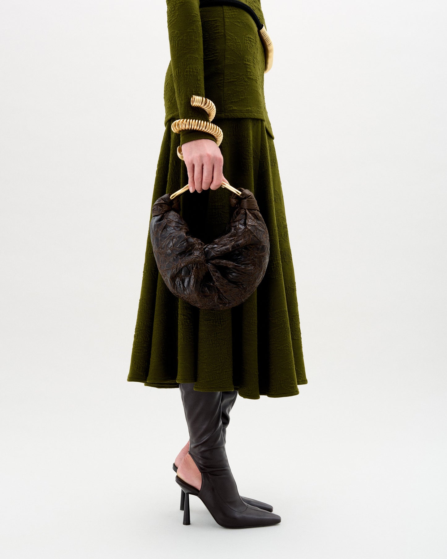 Una persona lleva un vestido verde oliva texturizado, botas negras de tacón alto, joyas doradas y el bolso Sofía (pequeño), un bolso de mano marrón oscuro con forma de media luna y asa redondeada, sobre un fondo liso.