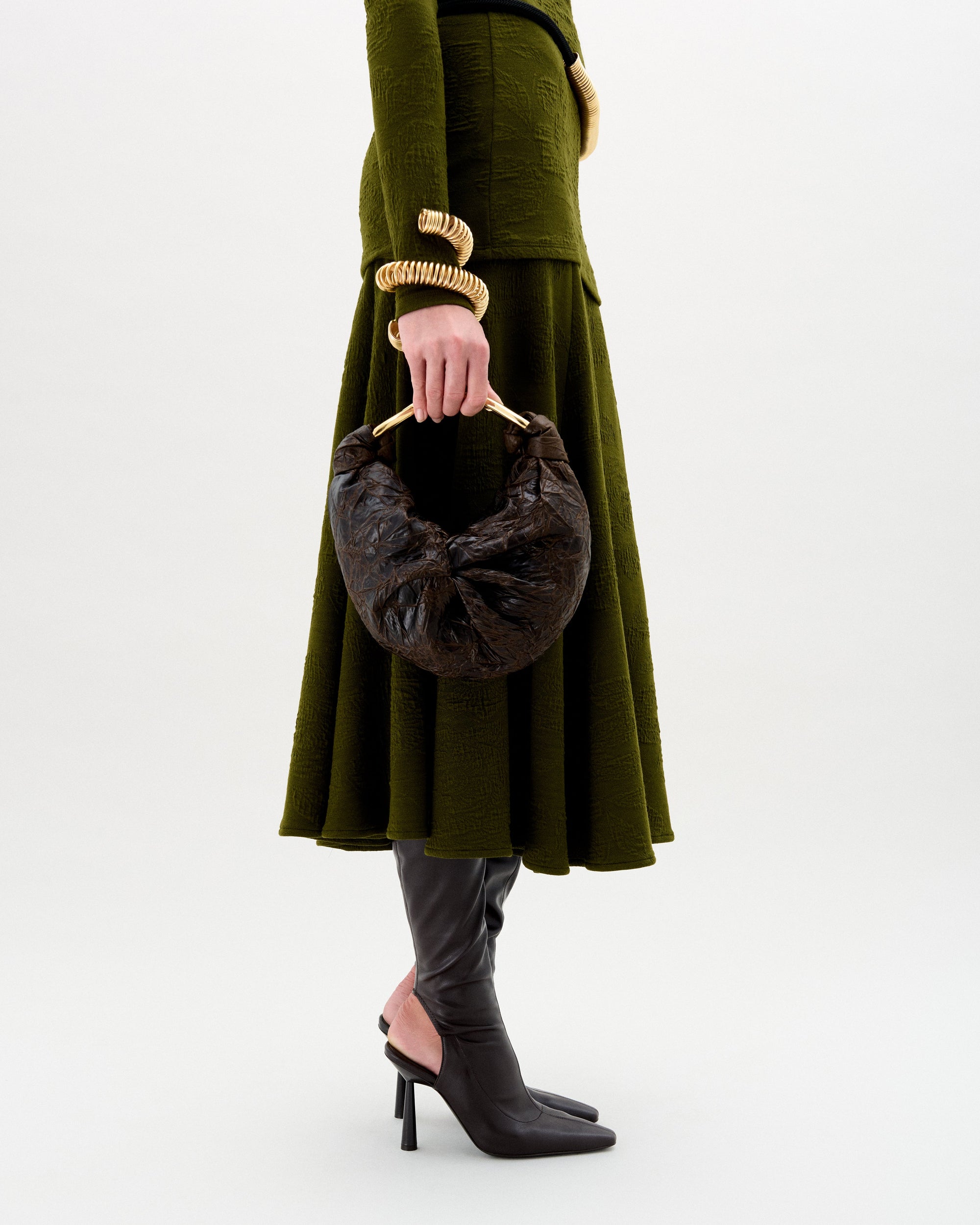 Una persona lleva un vestido verde oliva texturizado, botas negras de tacón alto, joyas doradas y el bolso Sofía (pequeño), un bolso de mano marrón oscuro con forma de media luna y asa redondeada, sobre un fondo liso.