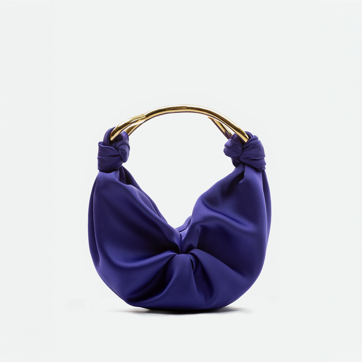 El Bolso Sofia Small es una cartera pequeña de color púrpura confeccionada en tela italiana, con un diseño suave y fruncido y un asa dorada curvada con extremos anudados a ambos lados. Hecho en Colombia. Sobre fondo blanco liso.