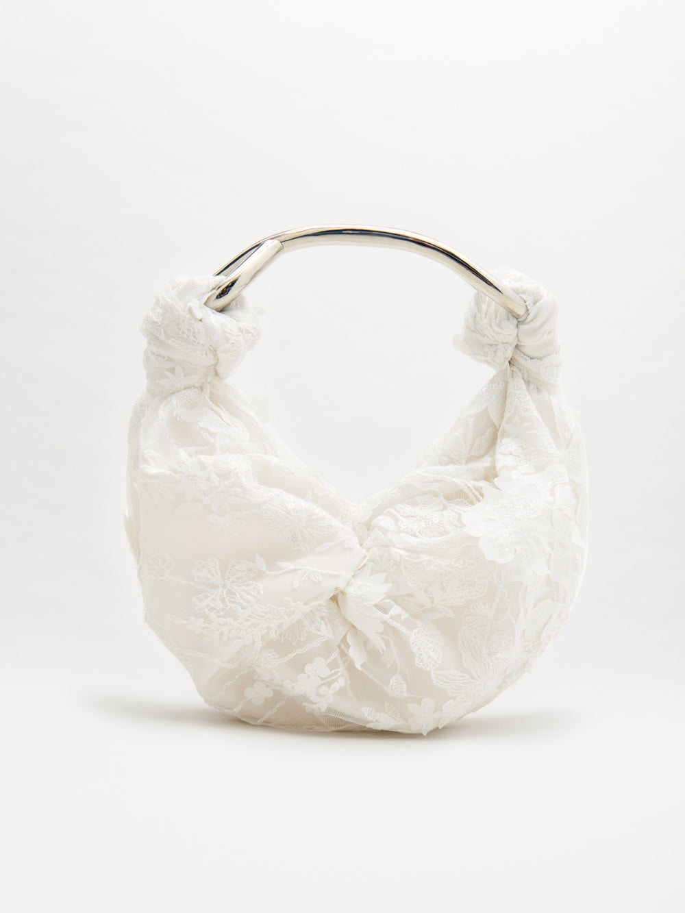El Bolso Sofía es un bolso blanco con una capa de encaje floral y un llamativo nudo en la base de su asa curva de metal plateado. Se presenta sobre un fondo blanco liso.