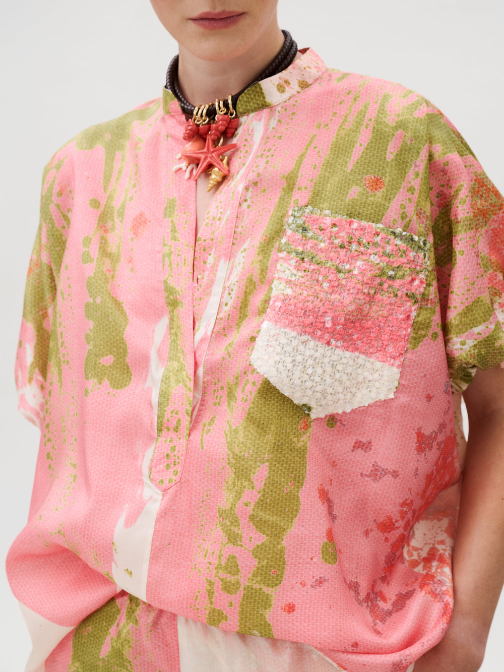 Susane Blouse in pink and green abstract silk habutai with a mandarin collar, short sleeves, sequined chest pocket, and a beaded seashell necklace.