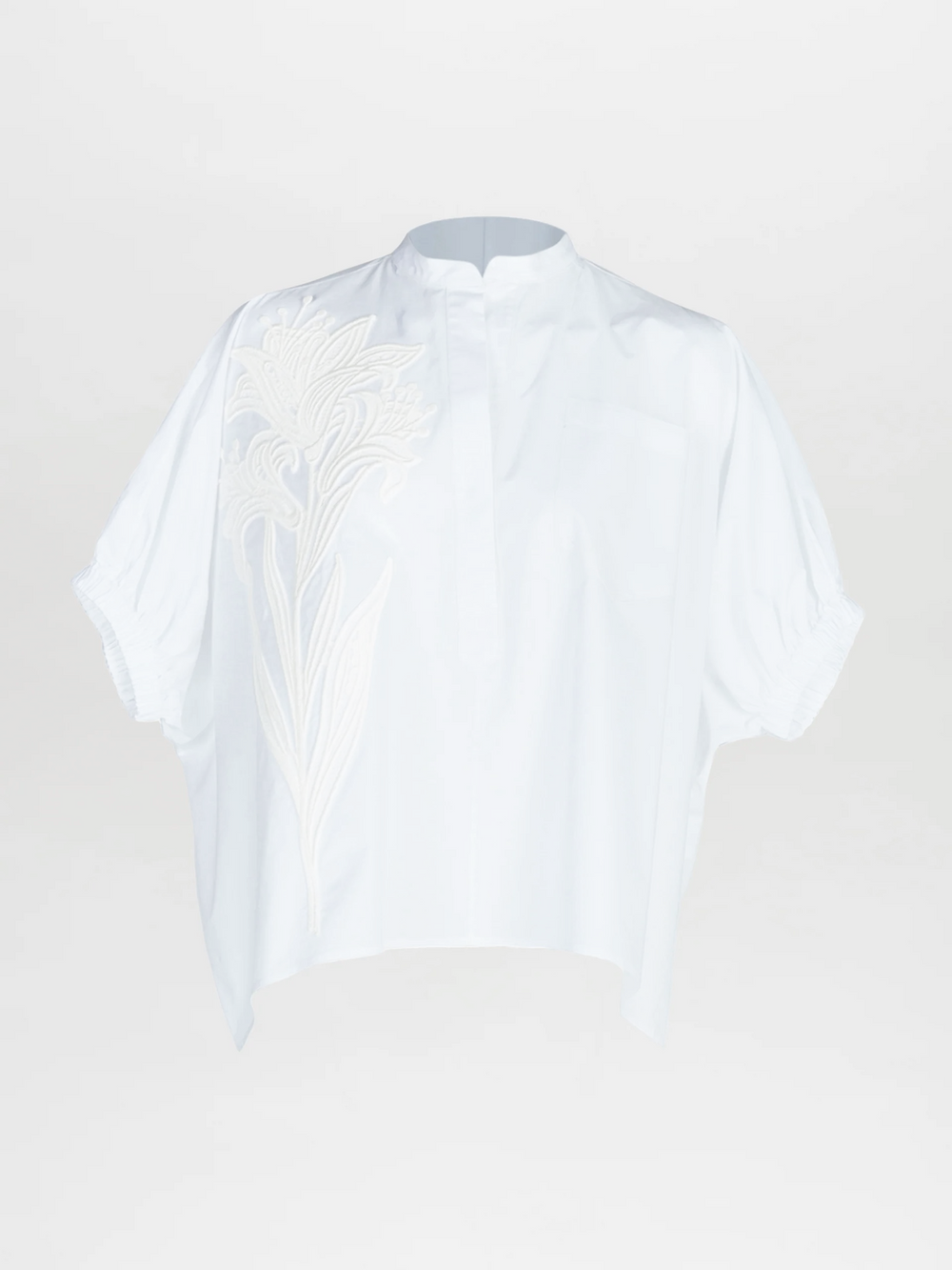 The Susanne Blouse is a loose-fitting, short-sleeved white top featuring a stand collar, subtle button placket, and oversized floral embroidery on the left shoulder and chest.