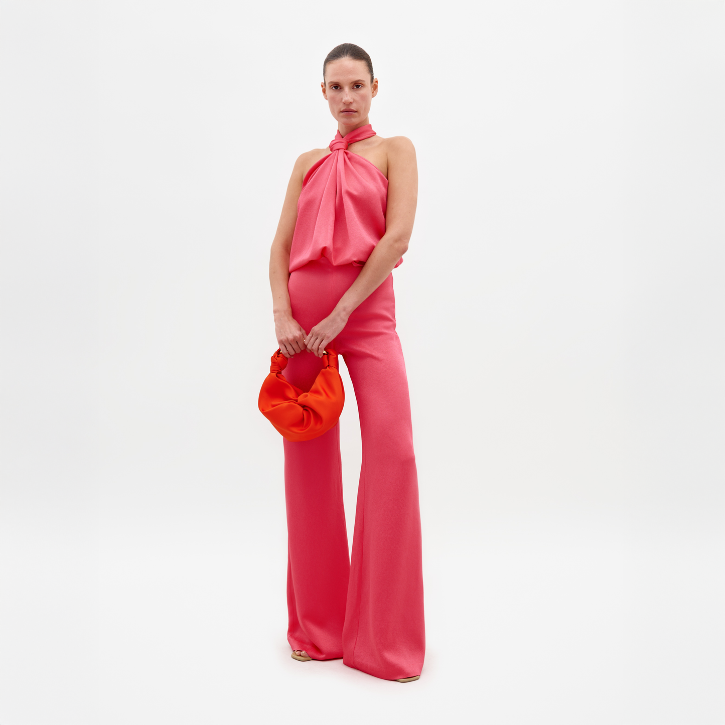 Sobre un fondo blanco liso, una persona luce con confianza un top rosa brillante con cuello halter y pantalones anchos Pantalón Palermo a juego, confeccionados en una exquisita tela italiana. Sostiene un bolso naranja, mostrando elegancia y estilo.