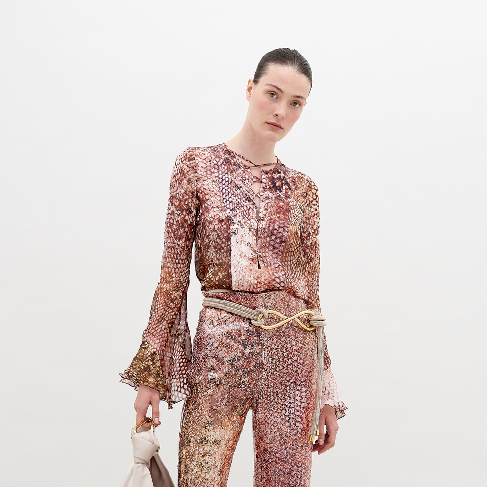 Sobre un fondo blanco, una persona lleva la Blusa Thea, una blusa de chifón de manga larga con estampado de serpiente combinada con pantalones y un cinturón decorativo con detalles metálicos. Sostiene un bolso de mano de color claro, lleva el pelo recogido hacia atrás y el tejido 100% seda del conjunto destila elegancia.