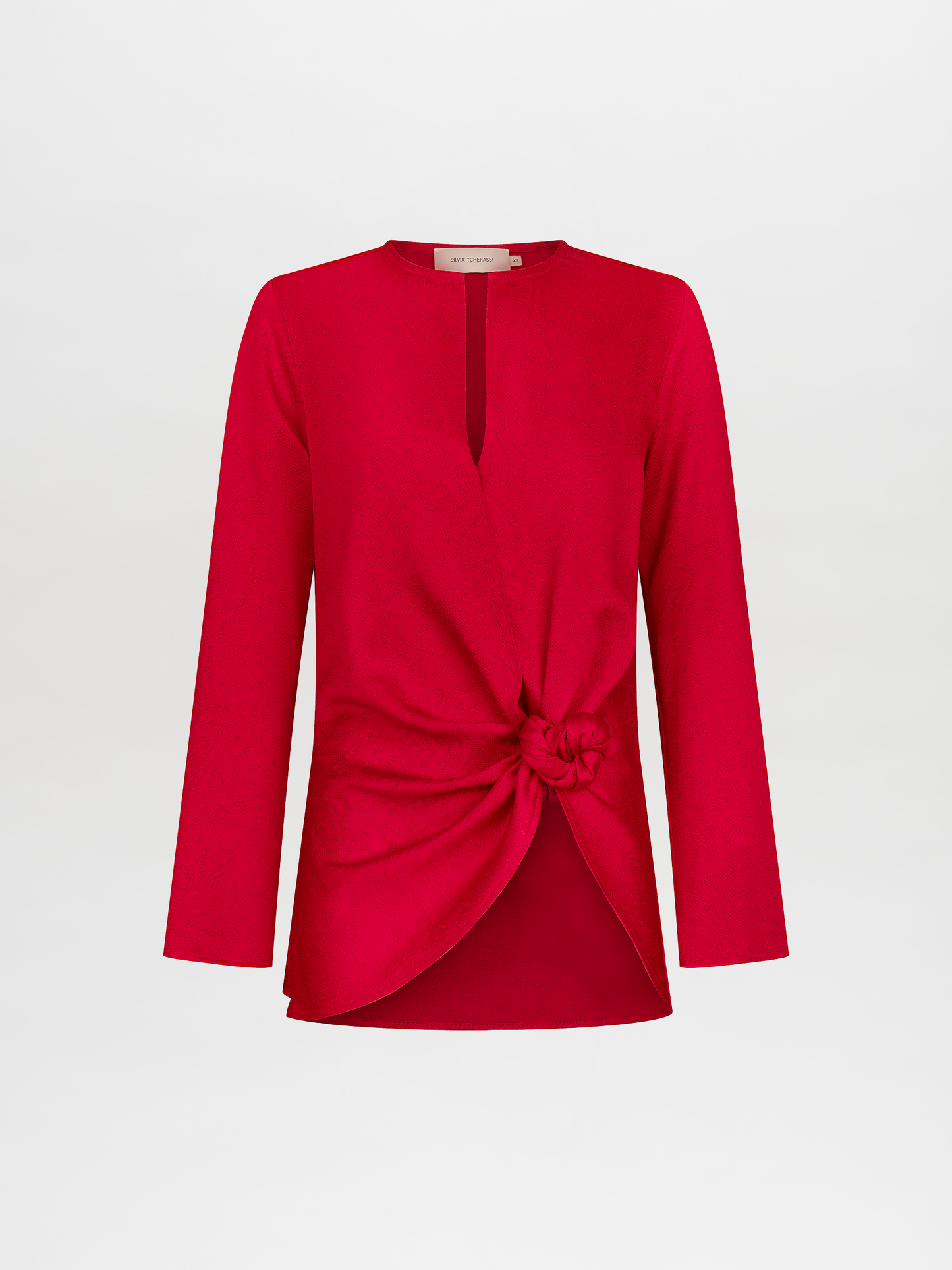 A woman stands in a red outfit with a knot detail on the top. She has her hair pulled back and stares directly into the camera against a plain white background. Available for pre-order now, the Tayra Blouse Rouge will have a ship date of November 15th, 2024.