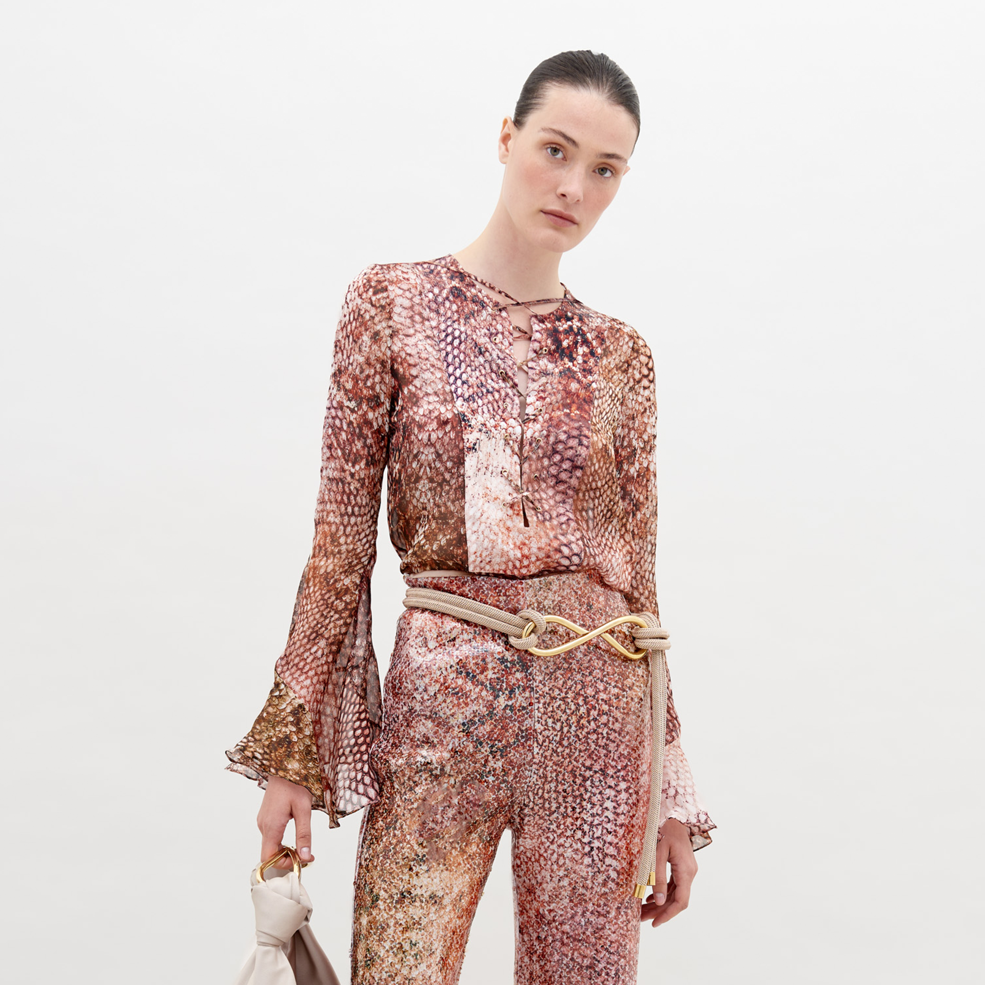 Una persona con el pelo recogido lleva la Blusa Thea, una blusa de gasa de manga larga con estampado de serpiente, puños acampanados y pantalones a juego. Lleva un cinturón de cuerda anudado y un bolso color crema sobre fondo blanco.