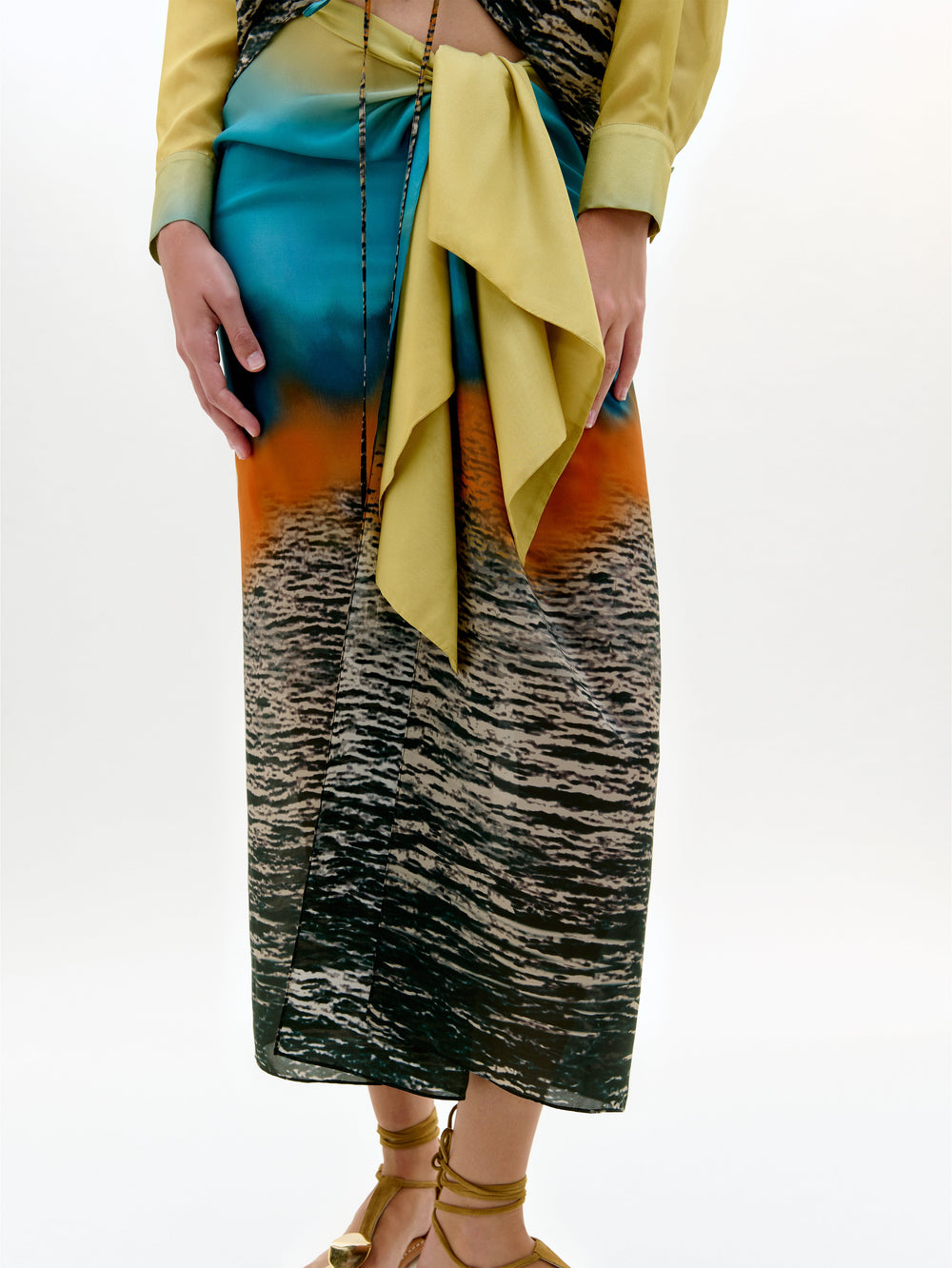 Una persona aparece sobre un fondo claro liso llevando el pareo Viveka, una falda midi hecha en Colombia con un estampado multicolor de aguas del atardecer, combinado con una faja amarilla, un top amarillo y sandalias de tiras.