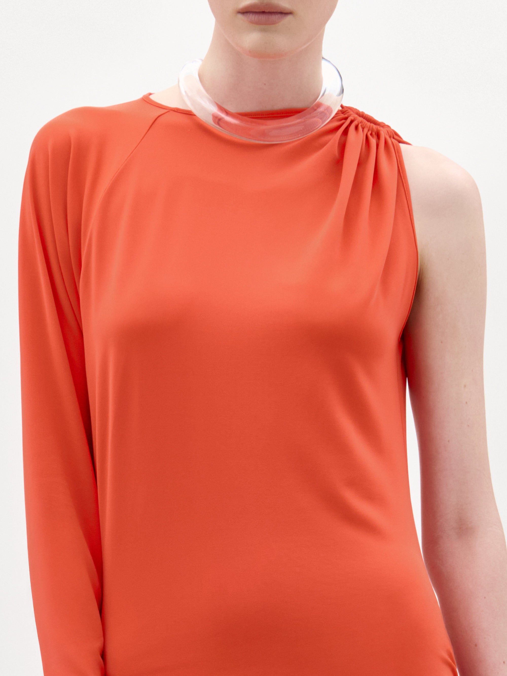 Una persona luce el vestido Virna: un maxivestido asimétrico naranja de una sola manga, combinado con un collar grueso transparente, sobre un fondo blanco liso. Este llamativo look está hecho en Colombia.