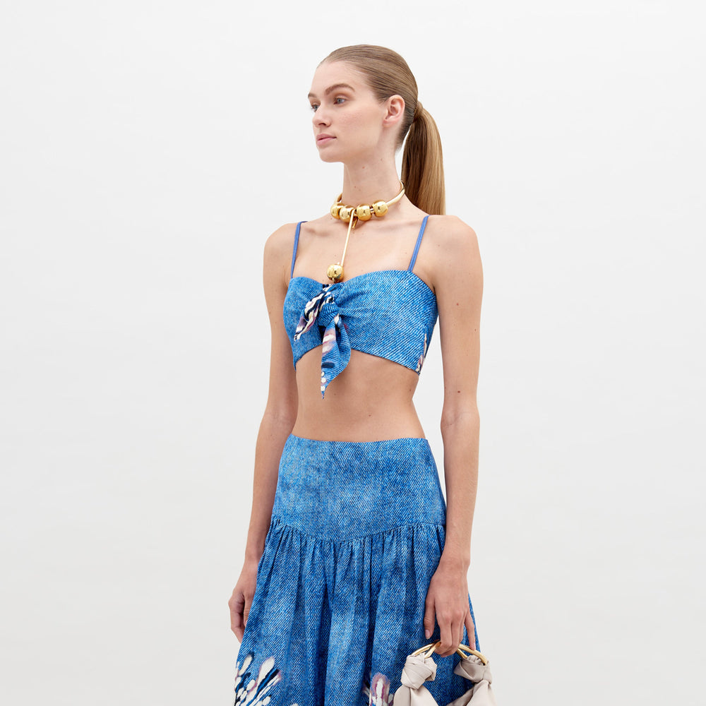 Una mujer con el pelo largo recogido en una cola de caballo lleva el Crop Top Wotton, un conjunto azul de dos piezas con un top corto con tirantes finos y un nudo en la parte delantera, posiblemente hecho en Colombia. Complementa su look con un collar dorado grueso y una pequeña cartera sobre un fondo blanco liso.