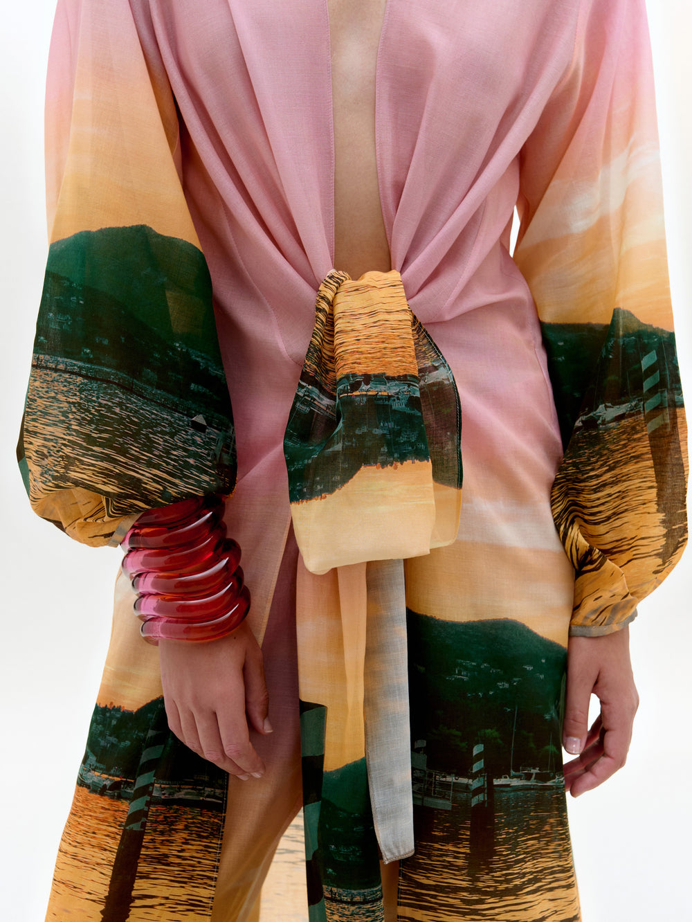 Una persona lleva la bata Yanil, una prenda de manga larga con estampado de paisajes en rosa y naranja, atada a la cintura. Confeccionada en algodón 100% con un profundo escote en V, se adorna con varios brazaletes de color rosa translúcido. La cabeza no es visible.