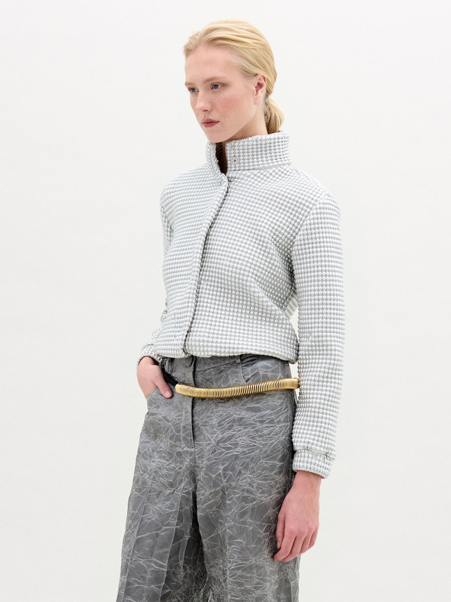 Una persona de pelo rubio lleva la chaqueta Zalta, una chaqueta corta de cuello alto y textura confeccionada en tejido italiano, combinada con pantalones grises arrugados y un cinturón ligero en forma de cuerda. Fabricada en Colombia. El fondo es blanco liso.