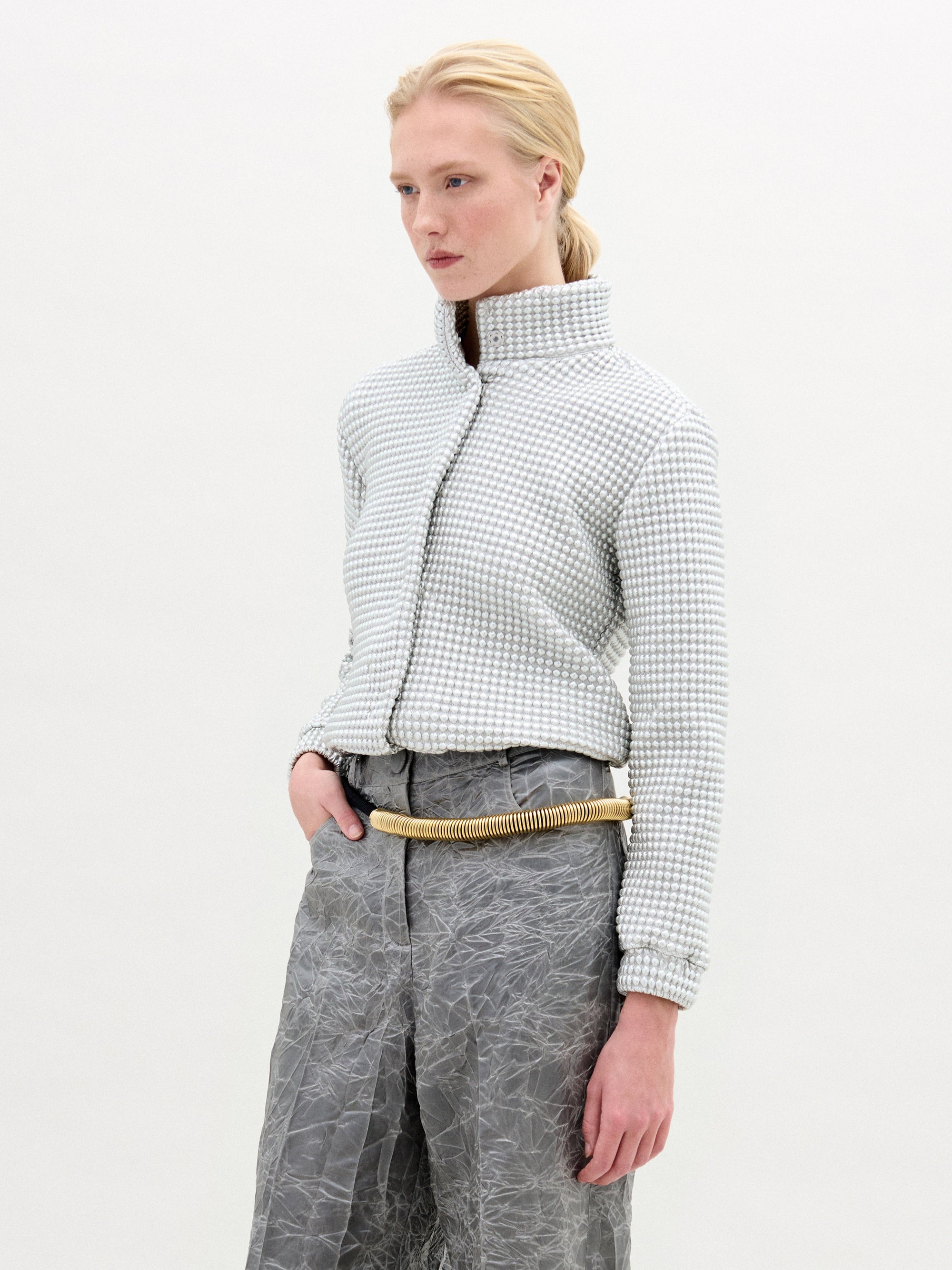 Una persona de pelo rubio lleva la chaqueta Zalta, una chaqueta corta de cuello alto y textura confeccionada en tejido italiano, combinada con pantalones grises arrugados y un cinturón ligero en forma de cuerda. Fabricada en Colombia. El fondo es blanco liso.