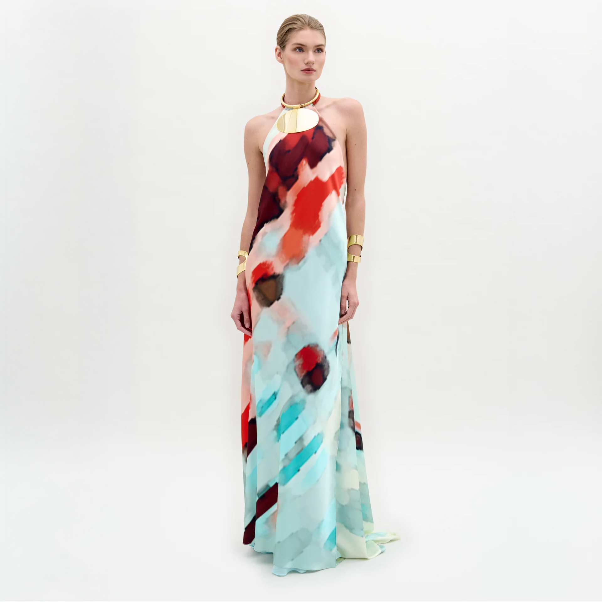 Una mujer sobre fondo blanco luce el vestido Zeffa, un maxi vestido de seda con cuello halter y estampado abstracto en rojo, azul y blanco. Lo combina con pulseras doradas y un grueso collar dorado.
