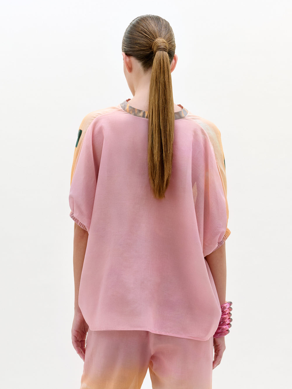 Una persona con el pelo castaño liso recogido en una coleta baja se ve de espaldas llevando la blusa Zenia, un top rosa suelto, translúcido y de manga corta con pantalones a juego, fabricado en Colombia. Lleva como accesorio una pulsera rosa gruesa. Fondo: blanco liso.
