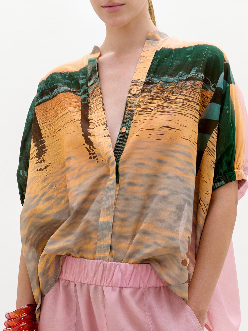 Lleva la blusa Zenia -una blusa holgada de manga corta con estampado de agua y atardeceres, escote en pico, botones y confeccionada en tela italiana- y la combina con unos pantalones rosas; su rostro queda parcialmente fuera del encuadre.