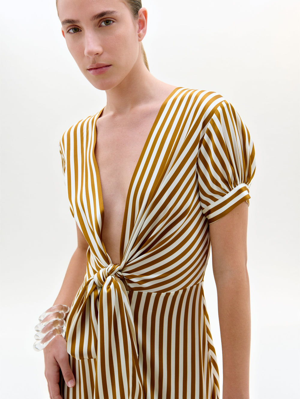 Una persona lleva el Vestido Zila -un midi de manga corta y escote en V con rayas verticales amarillas mostaza y blancas y un lazo delantero- y una pulsera transparente en la muñeca izquierda.