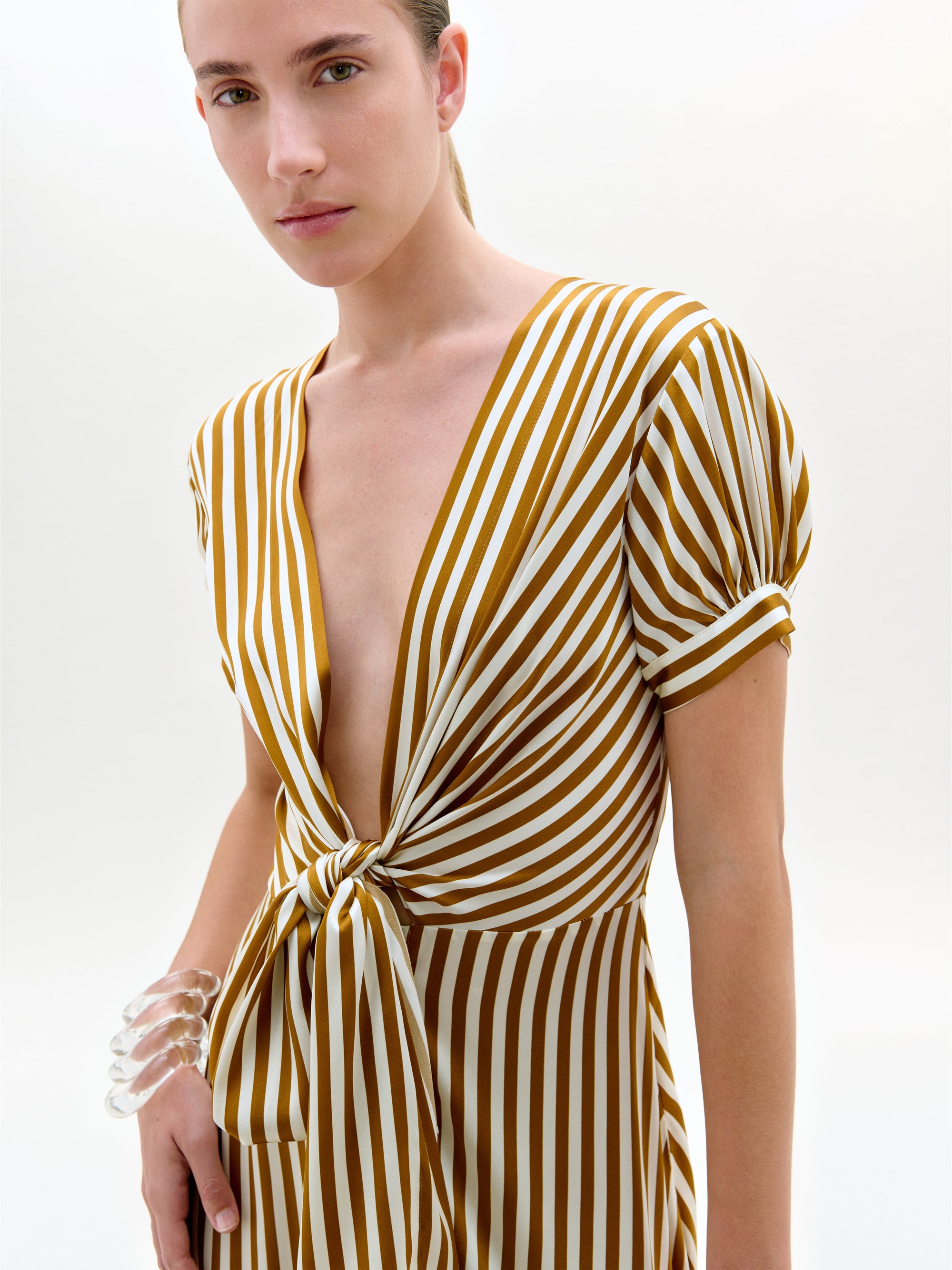 Una persona lleva el Vestido Zila -un midi de manga corta y escote en V con rayas verticales amarillas mostaza y blancas y un lazo delantero- y una pulsera transparente en la muñeca izquierda.