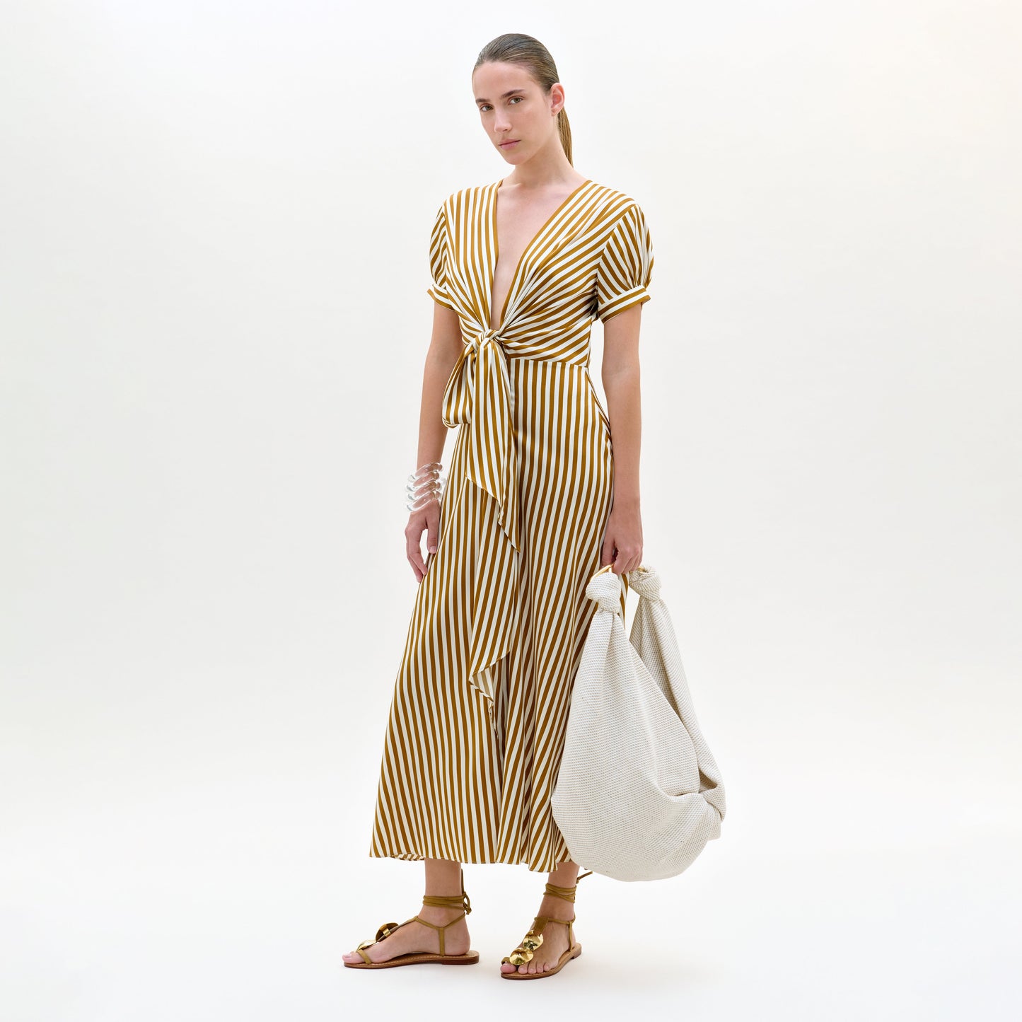 Una persona, sobre un fondo liso, lleva el vestido Zila, un vestido midi de seda a rayas verticales amarillas y blancas, con mangas cortas, escote en pico profundo y lazo delantero, combinado con sandalias planas y un bolso grande y ligero. Hecho en Colombia.