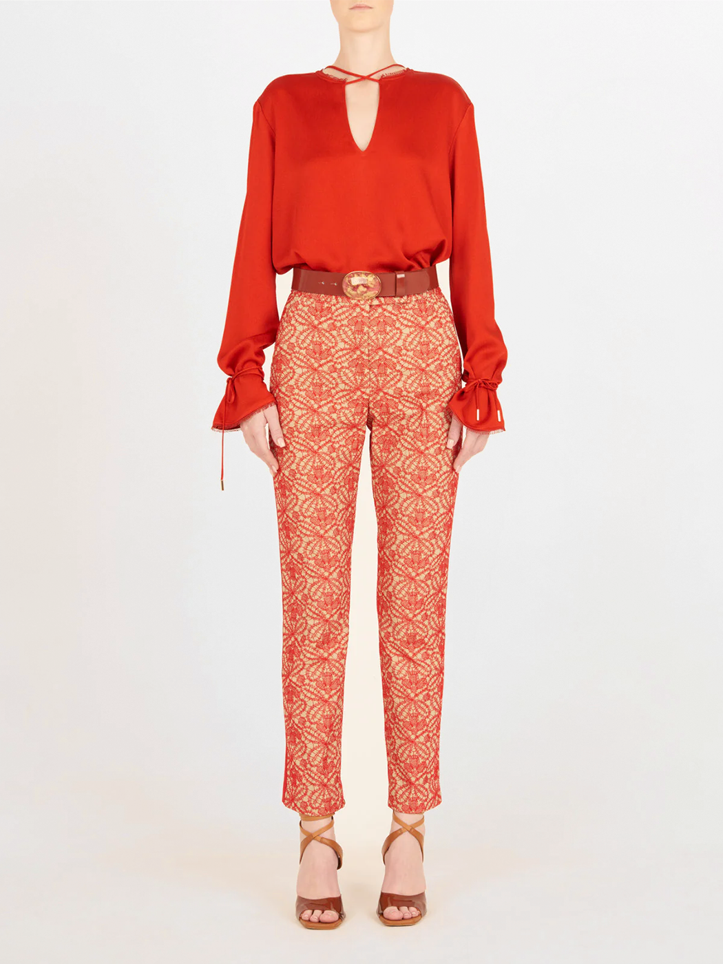 Una persona de pie sobre un fondo liso lleva un Blusa Zafra Rouge de manga larga, pantalones rojos y beige estampados, sandalias marrones de tacón alto y un cinturón marrón con hebilla redonda.