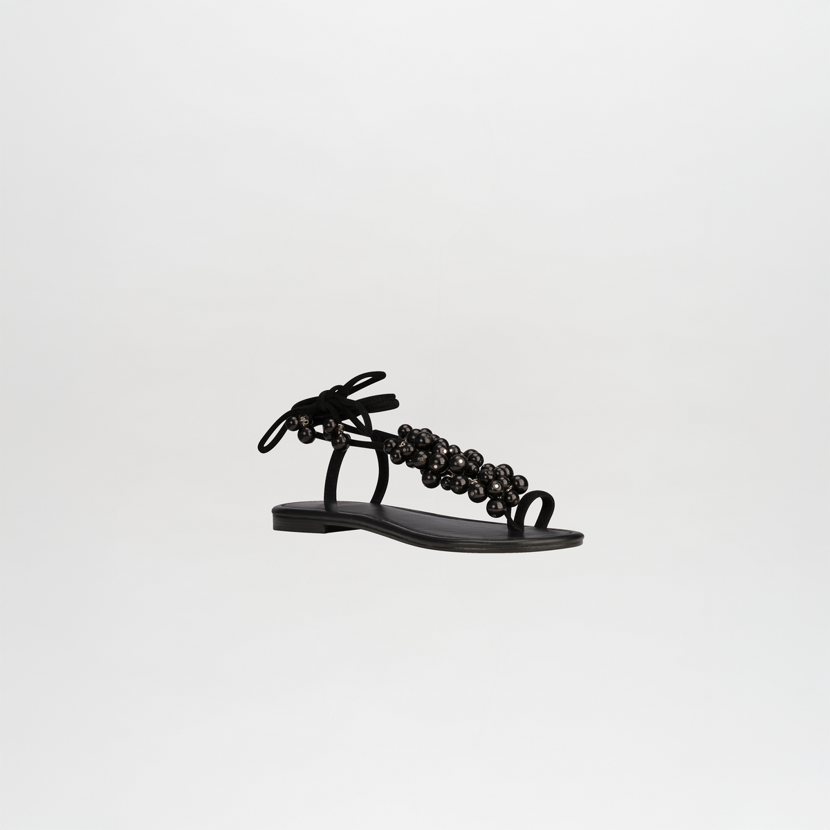 Las Sandalias Zaira, unas elegantes sandalias negras planas de cuero fino, presentan un diseño de tiras finas adornadas con elementos decorativos y se atan elegantemente alrededor del tobillo. Colocadas sobre un fondo blanco liso, destilan elegancia minimalista.
