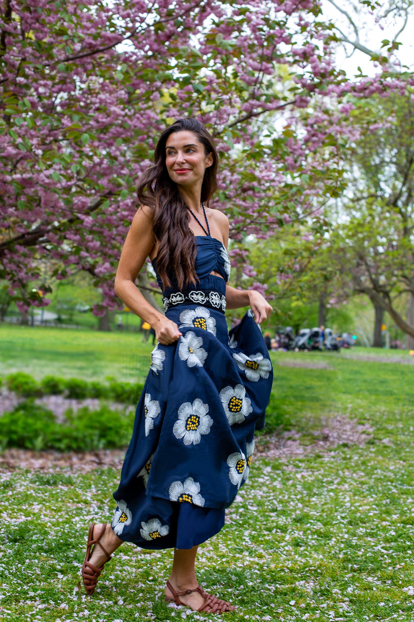 Una mujer con un vestido azul con estampados de flores blancas se encuentra en un parque con cerezos en flor. Sonríe y sostiene parte de su vestido. El suelo está cubierto de hierba verde y pétalos caídos. Lleva sandalias marrones.