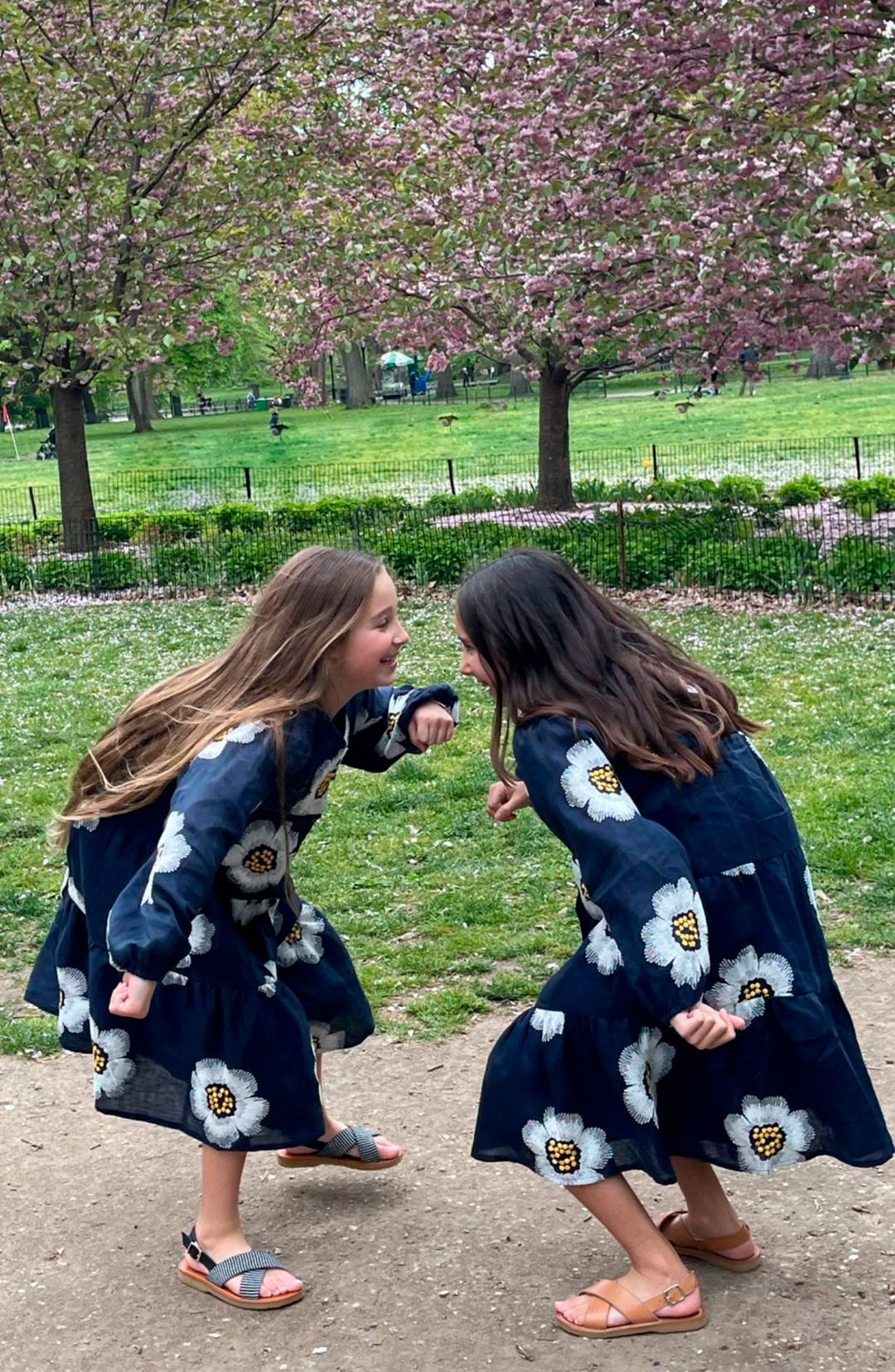 Dos niños se miran uno al otro al aire libre, llevan vestidos de color azul oscuro con estampados de flores blancas, se inclinan hacia delante de forma juguetona y parecen estar en un parque con árboles en flor al fondo.