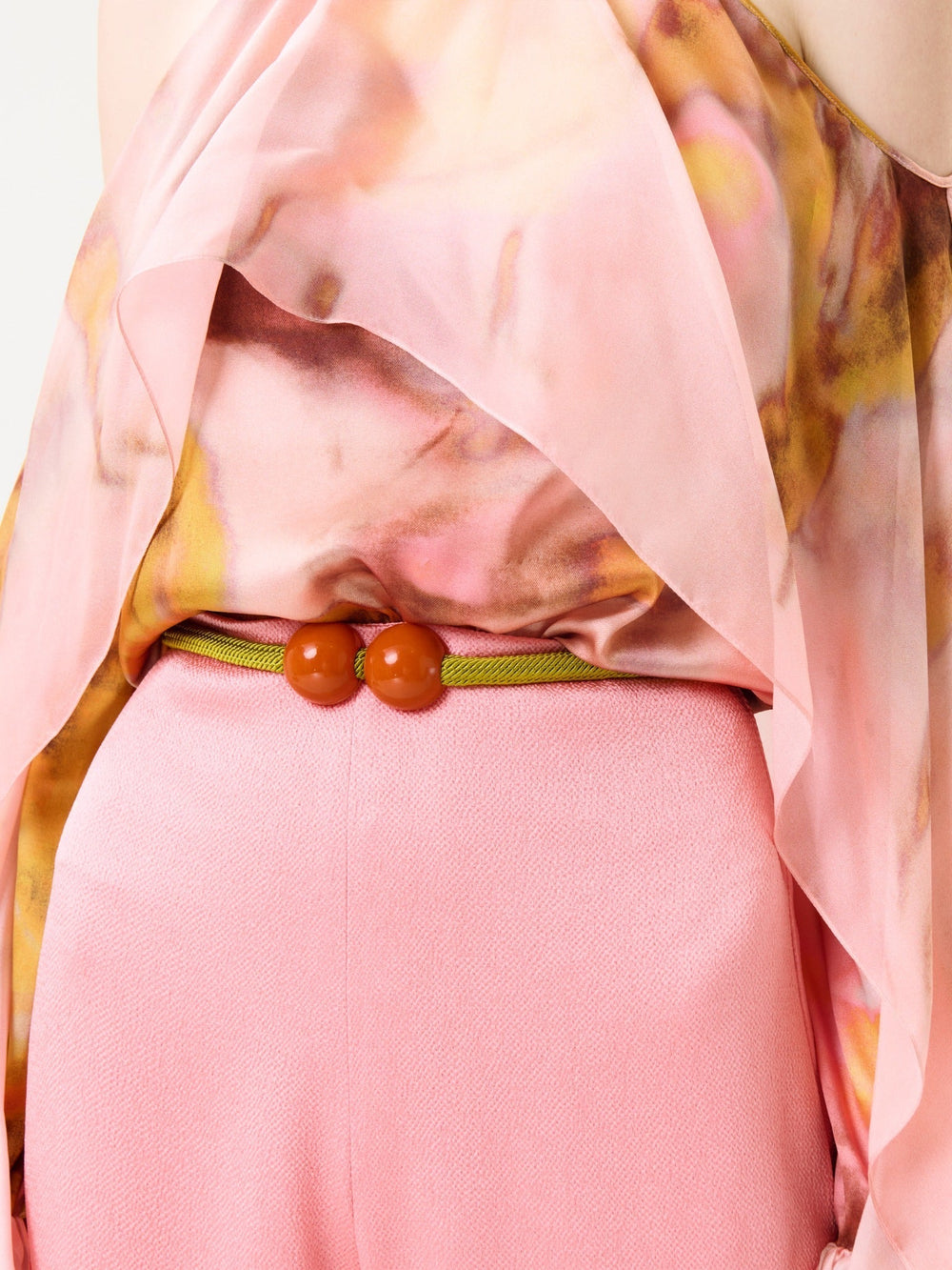 A person wearing the Shaan Blouse—a flowing, pink patterned top—paired with pink pants and a thin green belt featuring two round orange beads at the waist.
