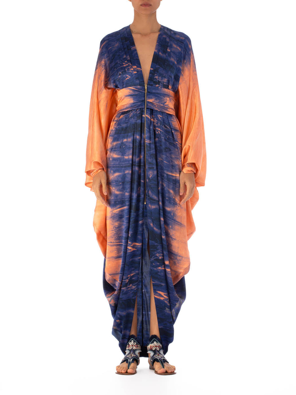 Una persona viste el elegante Vestido Danette Mediterranean Coral Blue, con un escote en V pronunciado y un estampado abstracto en azul y naranja. Las mangas largas y sueltas realzan el look, complementadas con sandalias planas con tiras sobre un fondo blanco liso.