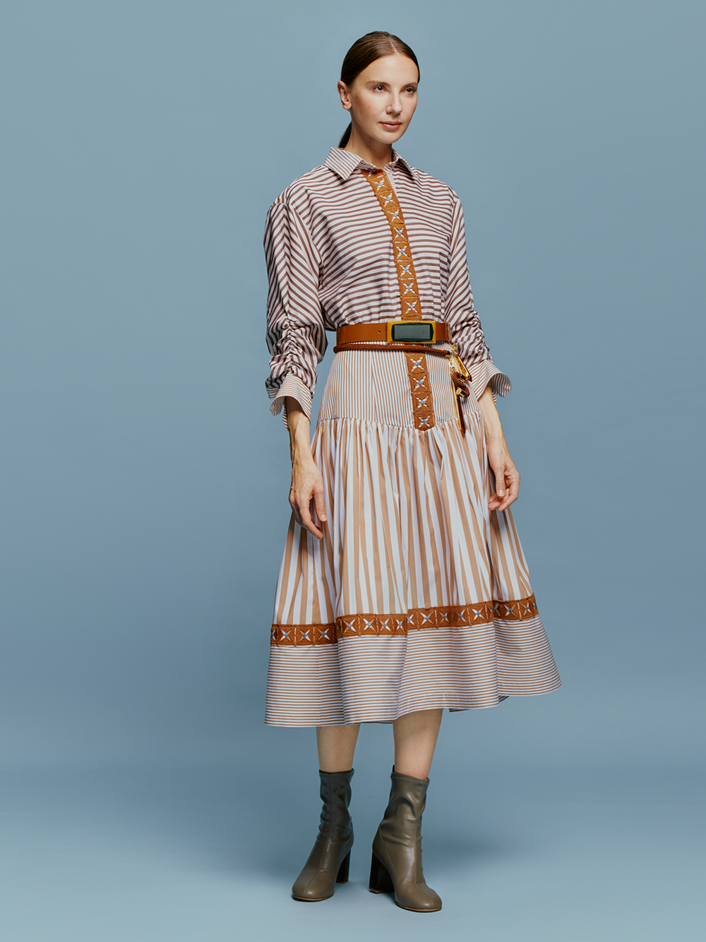 A woman stands against a blue background, wearing the Lamar Dress in Multi Brown Stripes, accentuated with a belt and boots, evoking the spirit of an August 19th summer day.