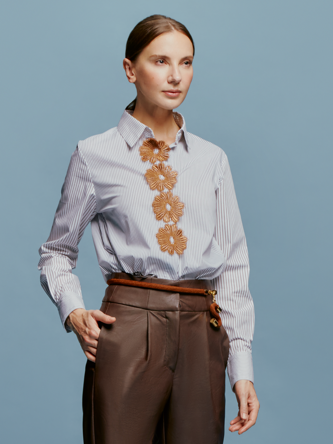 A brown-haired woman with her hair tied back is wearing the Naroa Blouse Multi Brown Stripes, featuring floral embroidery, along with brown pants. She is standing in front of a light blue background, indicating the date as August 19th, 2024.