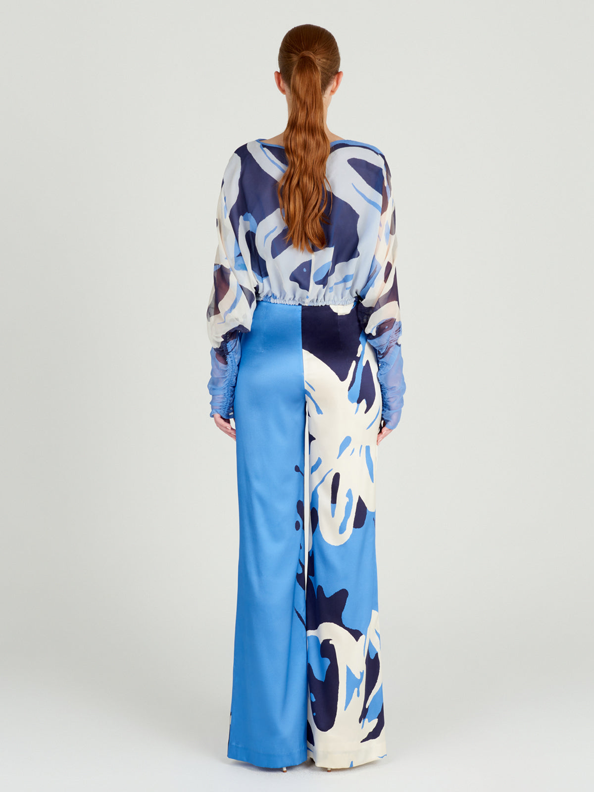 Una persona de pelo largo y pelirrojo está de espaldas, luciendo la blusa Macerata Celeste Bloom y unos pantalones anchos a juego. El conjunto luce un diseño de gasa transparente con un gran estampado abstracto en tonos azules y blancos que destila elegancia.