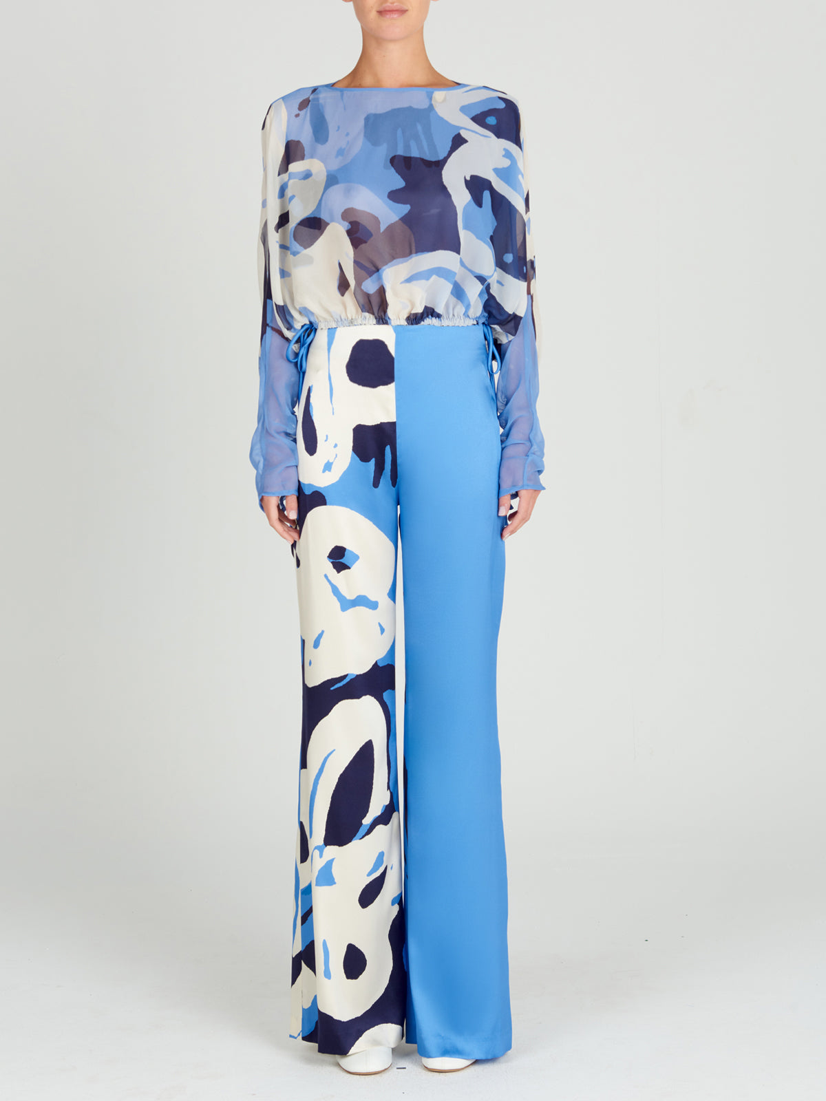 La persona lleva la blusa Macerata Celeste Bloom, con mangas largas y un estampado abstracto en azul y blanco. Los pantalones anchos tienen detalles en azul liso con patrones a juego. Un fondo liso resalta este llamativo conjunto de gasa mientras están de pie mirando hacia adelante.